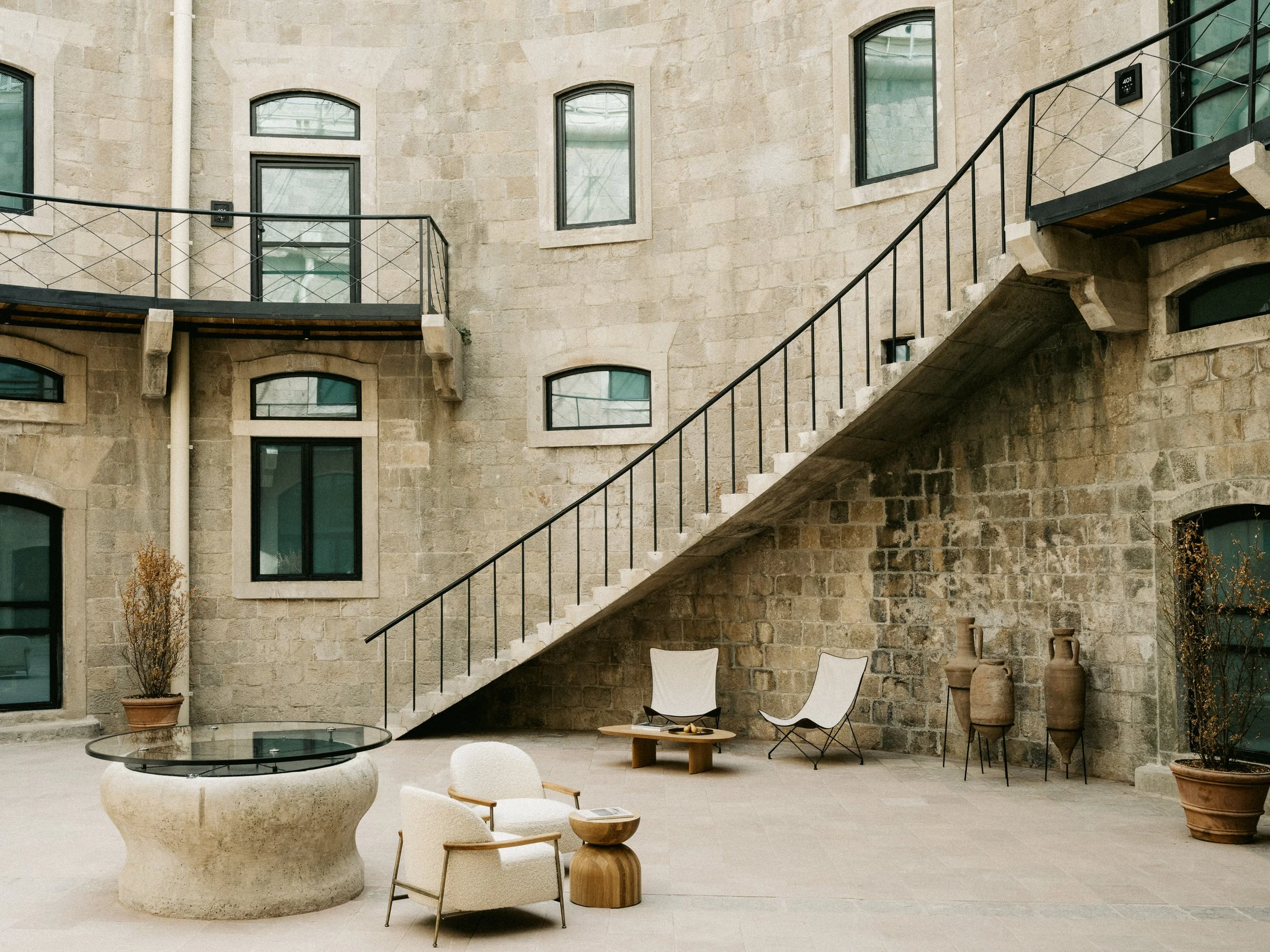 Interior courtyard of a stone building with multiple windows, outdoor staircase, potted plants, and modern furniture including chairs, a coffee table, and decorative objects.