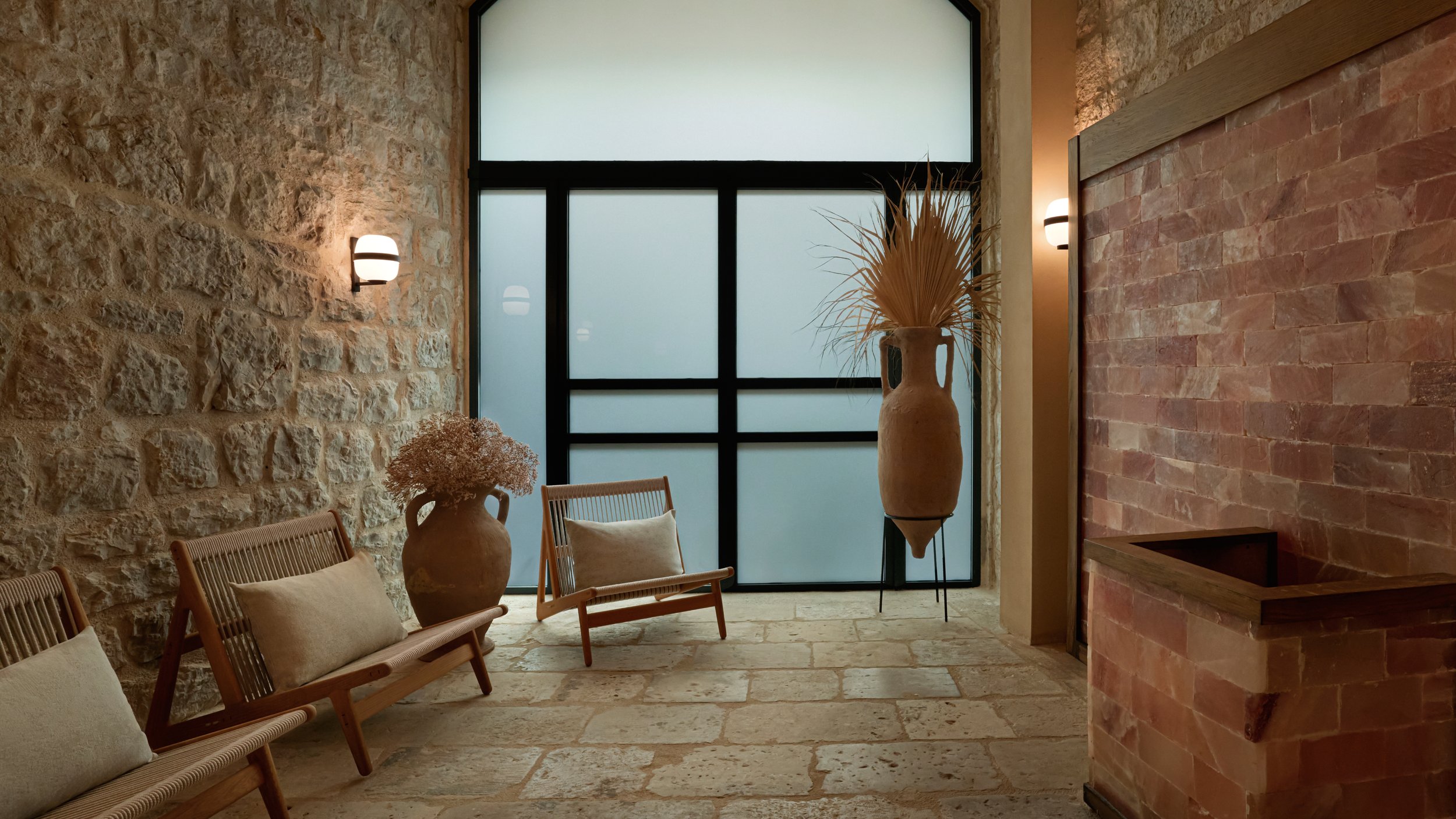 Interior of a room with stone and brick walls, a large window with frosted glass, decorative vases with dried plants, wooden chairs with cushions, and wall-mounted lights.
