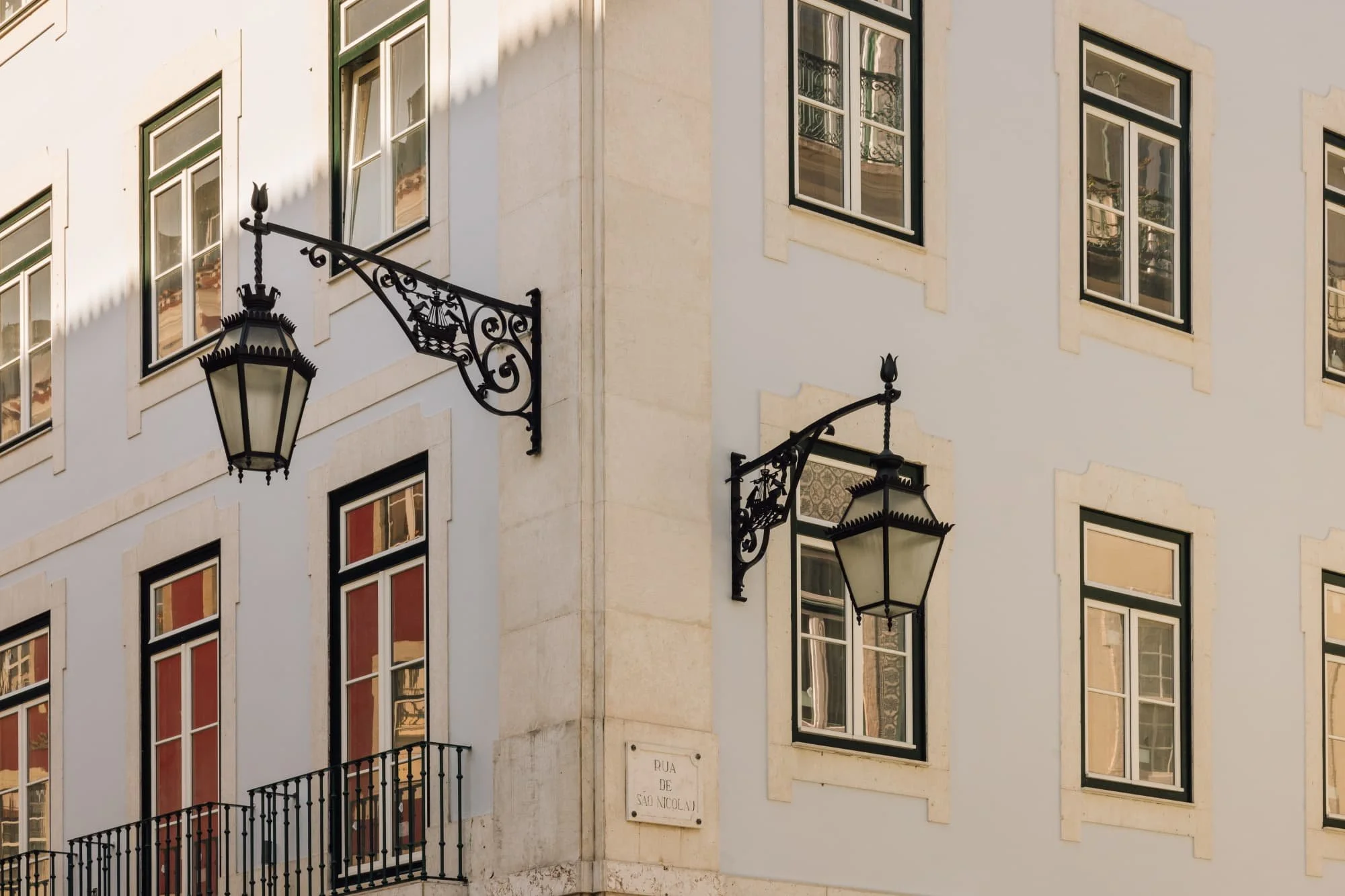 The image shows a corner of a white building with multiple windows, decorative iron street lamps, and a street sign that reads 'Rua de São Nicolau'.