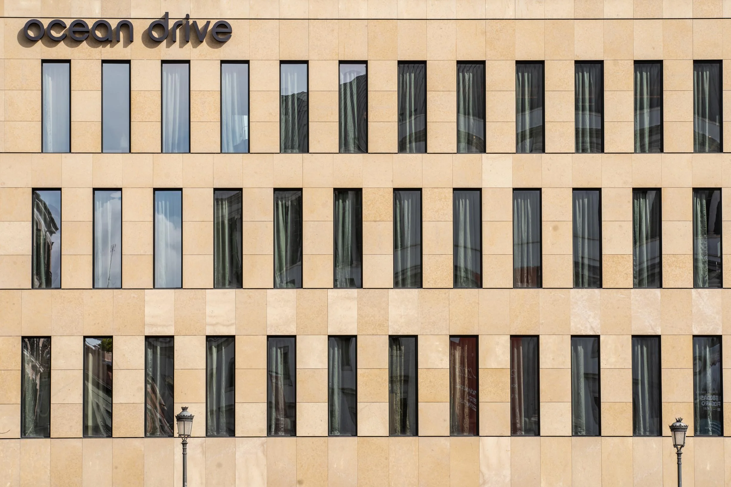 Exterior view of a beige building with the words 'ocean drive' on the upper left. Multiple vertical windows with curtains and reflections. Two street lamps are visible in the scene.