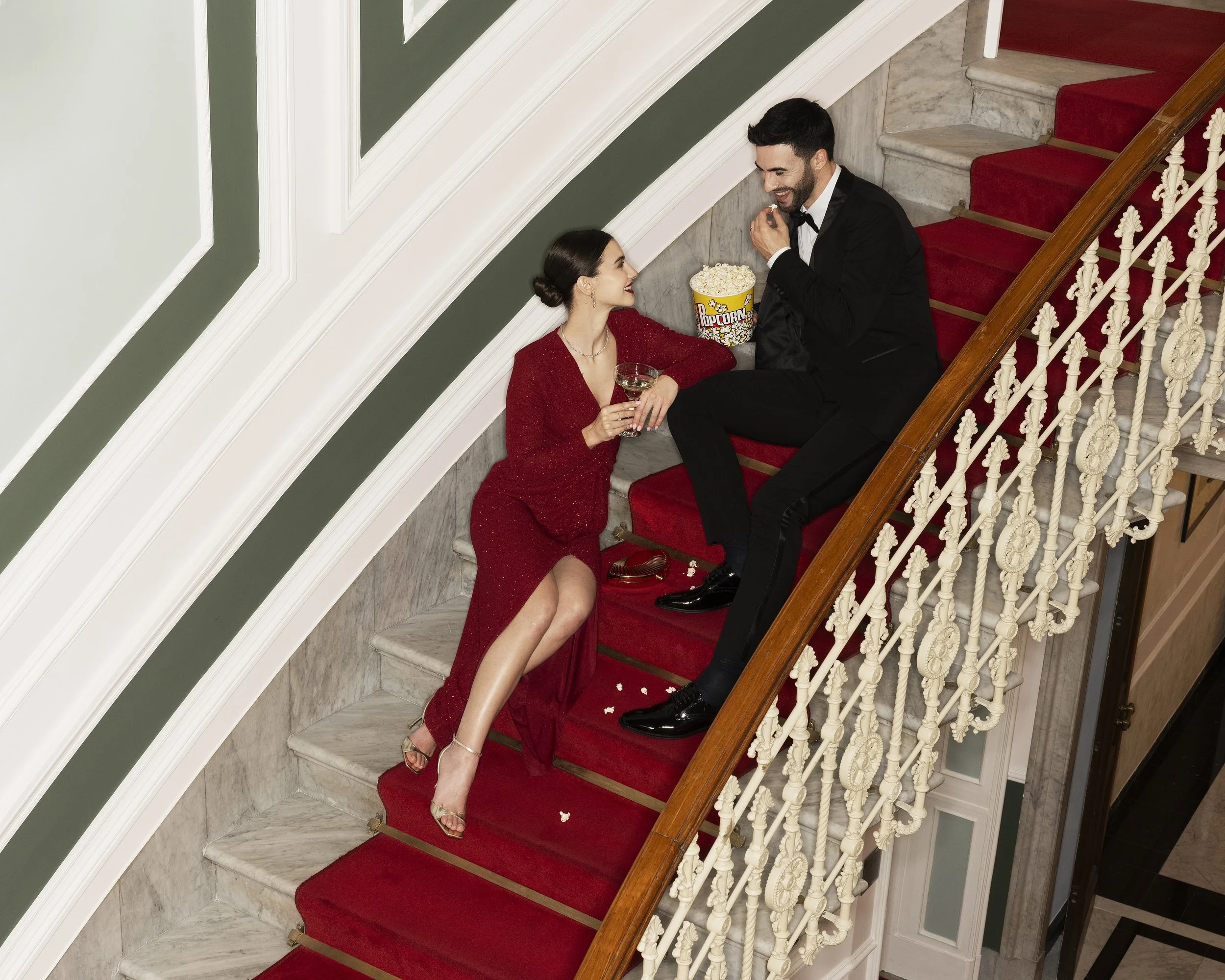 A man and woman sitting on a staircase at a formal event. The woman is wearing a red dress and holding a glass of champagne, and the man is wearing a black tuxedo and eating popcorn from a bucket.