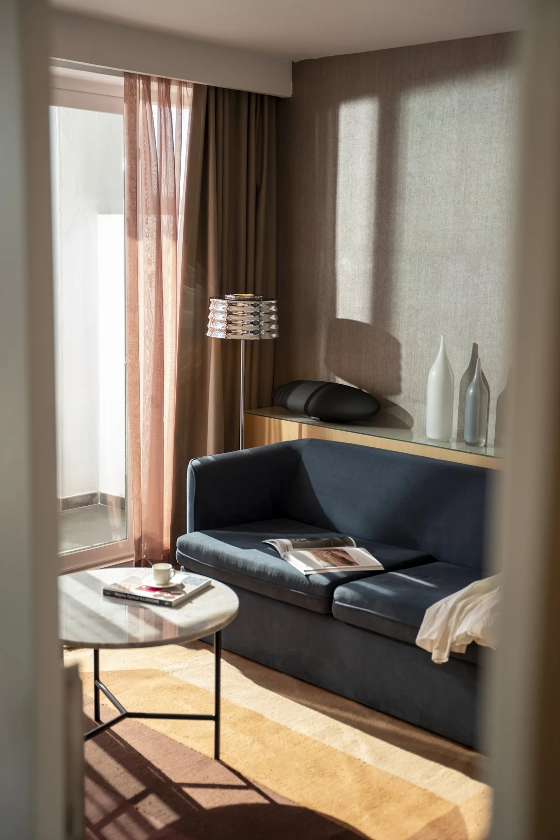 Living room with a navy sofa, round side table with a cup and magazine, beige carpet, large window with sheer and blackout curtains, floor lamp, decorative vases on a shelf, lit by natural sunlight.