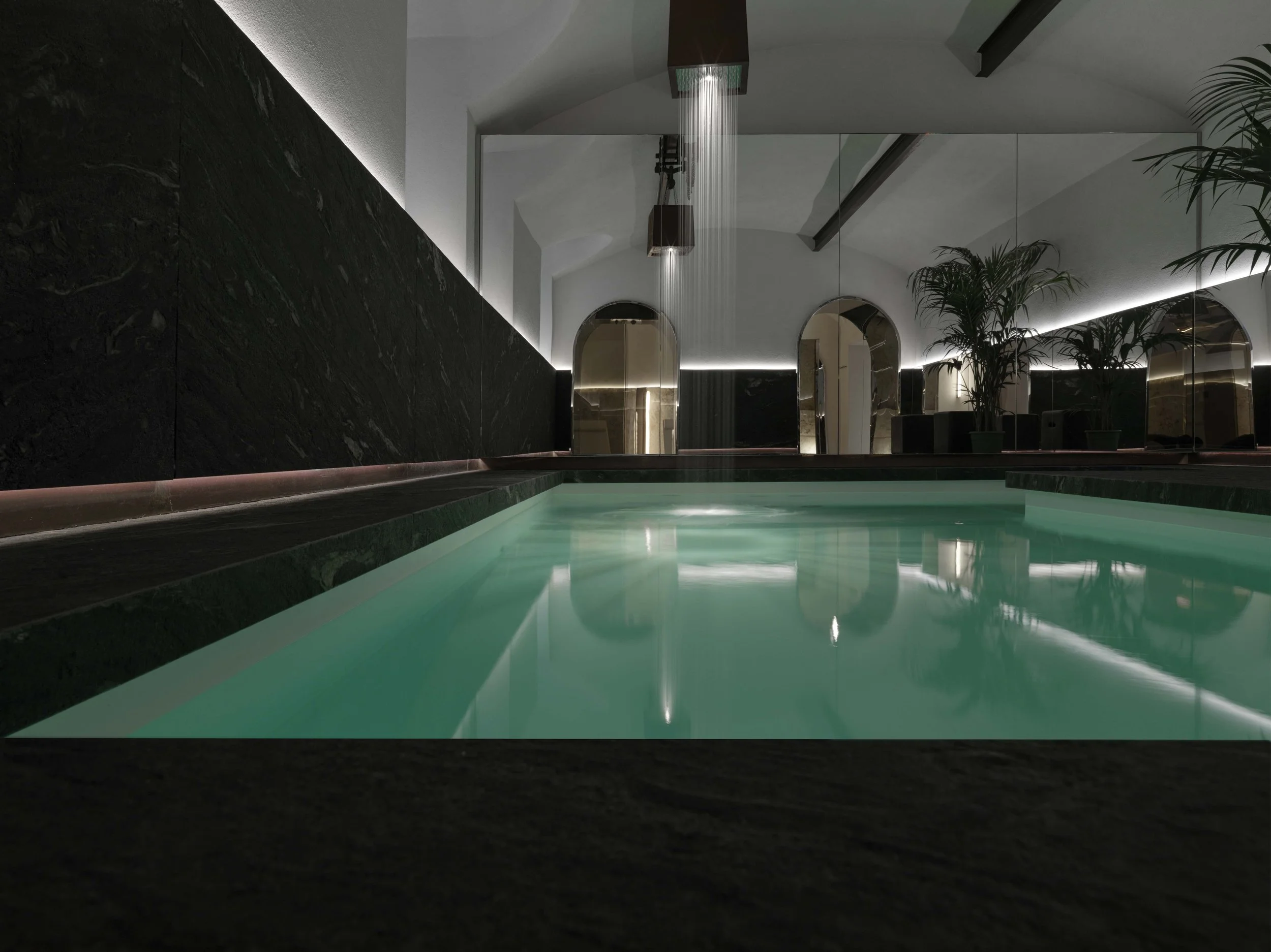 Indoor pool with-lit water, black stone wall, large mirrors, potted plants, and minimalist lighting fixtures.
