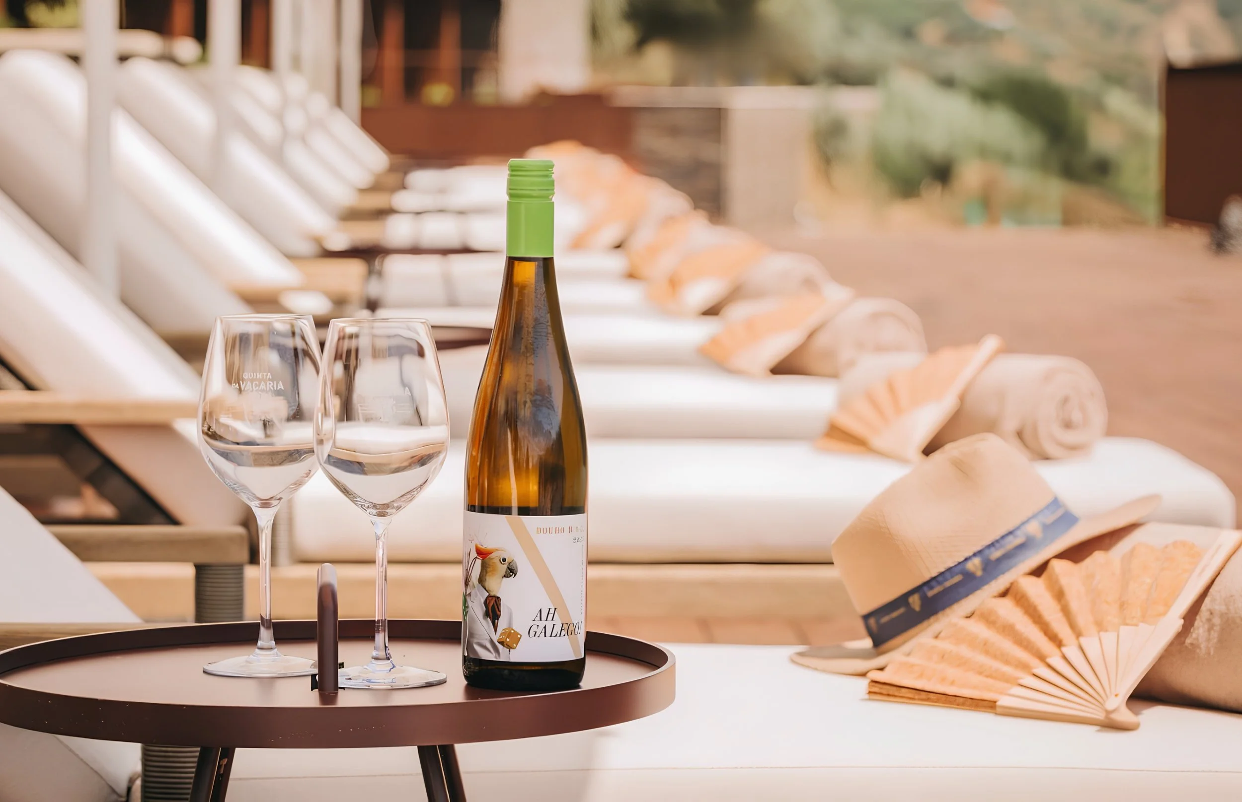 A bottle of wine and two wine glasses on a round table with a tray, hats, and paper-folded fans in the background on a patio or deck.