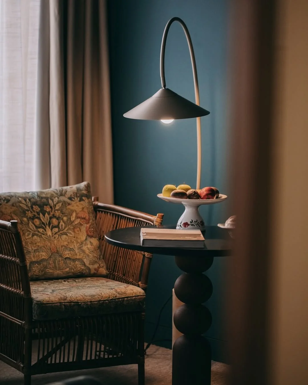 Table lamp casting light on a round table with a bowl of apples and a book, next to a floral cushion on a wicker chair in a cozy room with teal walls and beige curtains.