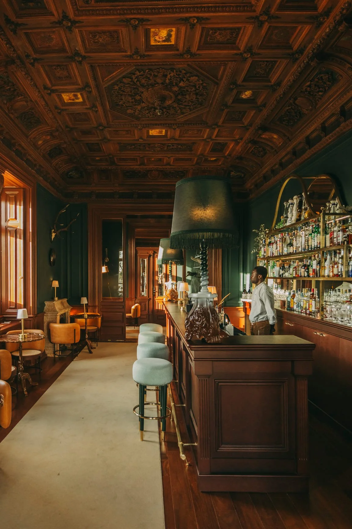 A luxurious bar interior with dark wood paneling, ornate ceiling, and large green lamps. Bar staff stands behind a polished wooden counter with a well-stocked liquor shelf. There are several cushioned chairs and small tables along the walls, with warm, ambient lighting.