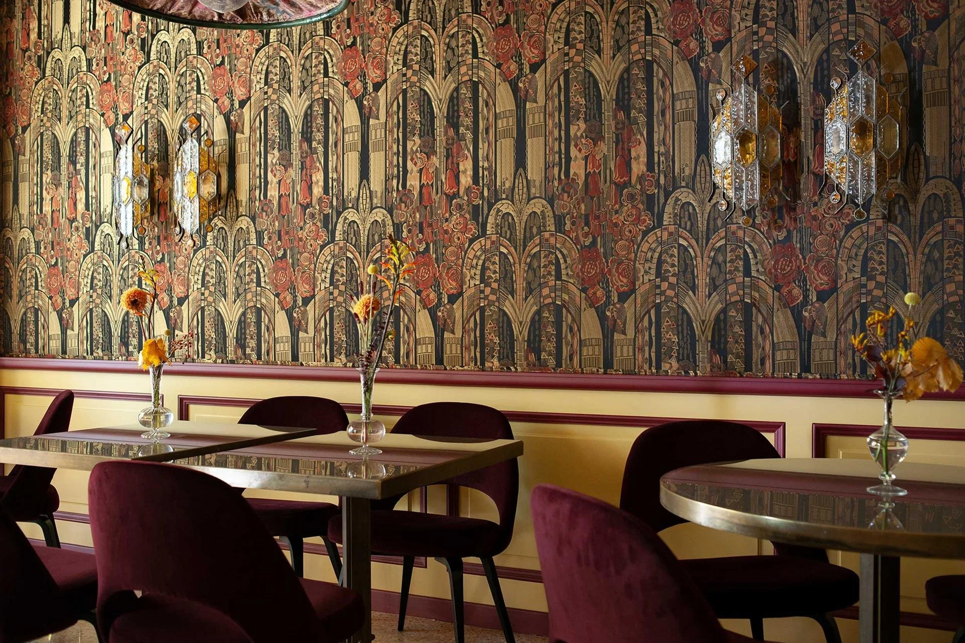 Interior of a dining area with a decorated wall featuring arched patterns and floral designs, four tall vases with flowers on tables, and dark red velvet chairs.