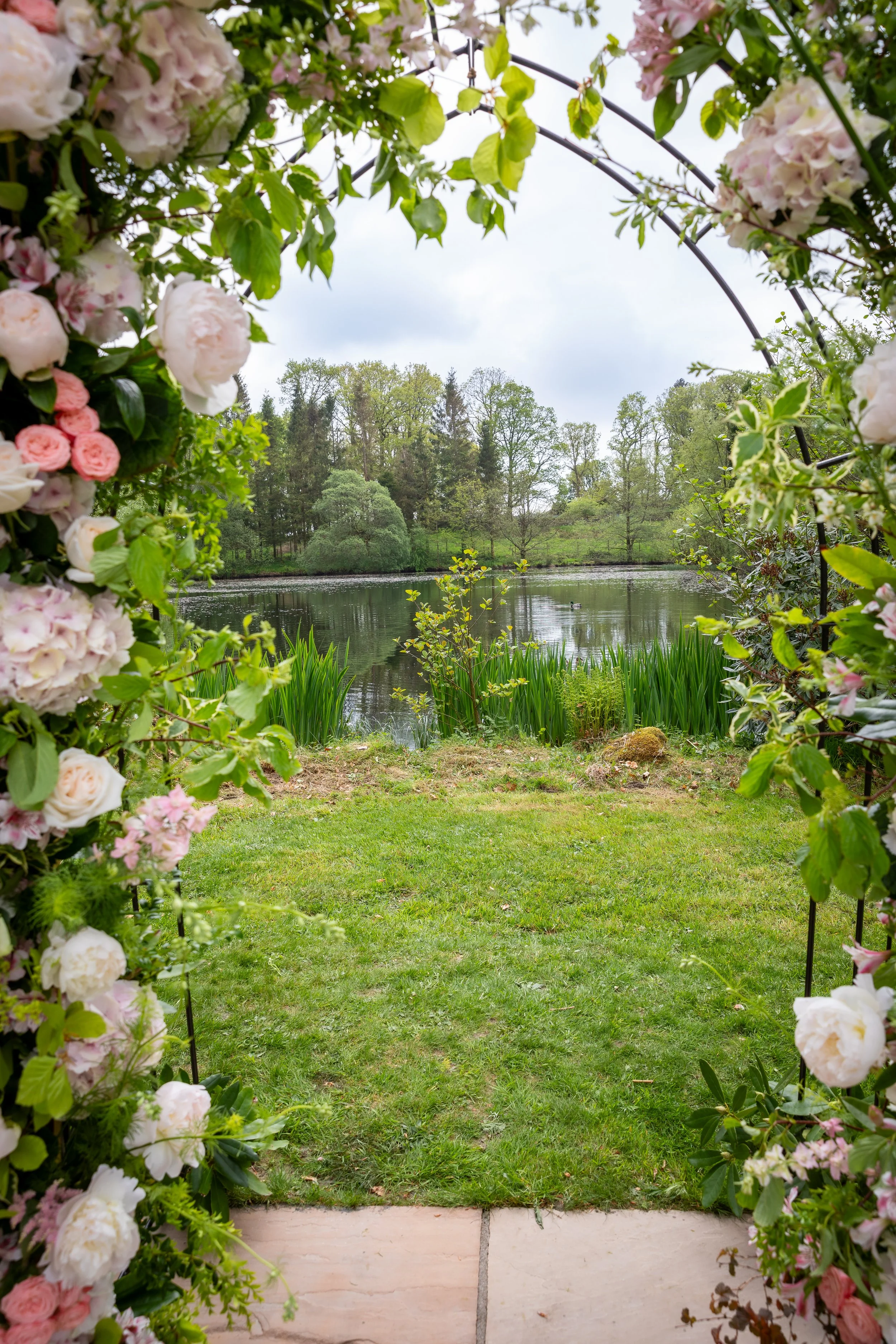 View of a lake surrounded by trees with an archway made of pink and white flowers and green leaves in the foreground.