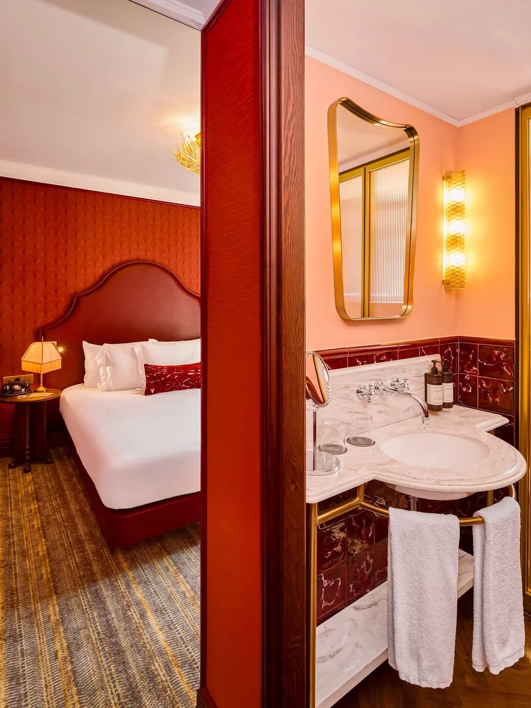 Split view of a hotel room with a bedroom on the left and a bathroom on the right. The bedroom has a bed with a tufted headboard, a nightstand with a lamp, and warm lighting. The bathroom features a marble sink with gold fixtures, a large mirror, and warm pink walls.