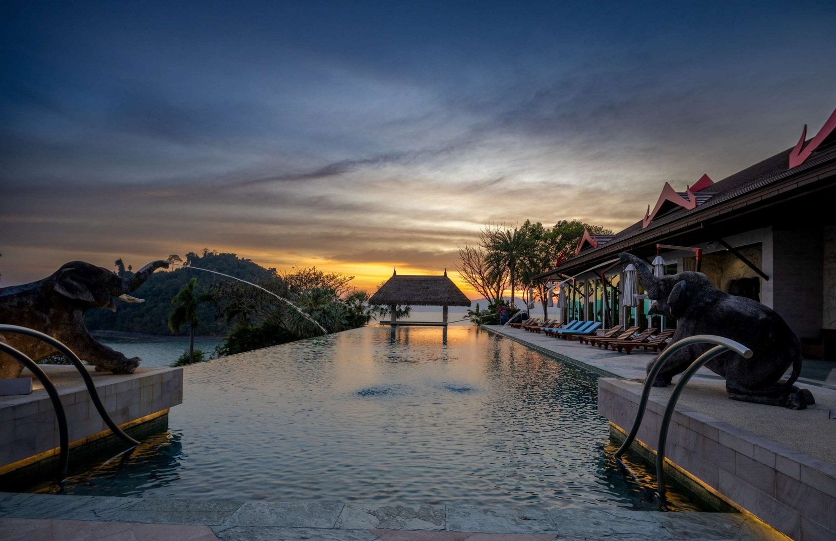 A luxurious infinity pool at sunset with decorative elephant sculptures on the sides, overlooking a scenic landscape with hills and trees. There is a small covered structure at the far end of the pool and a building with a sloped roof and red accents