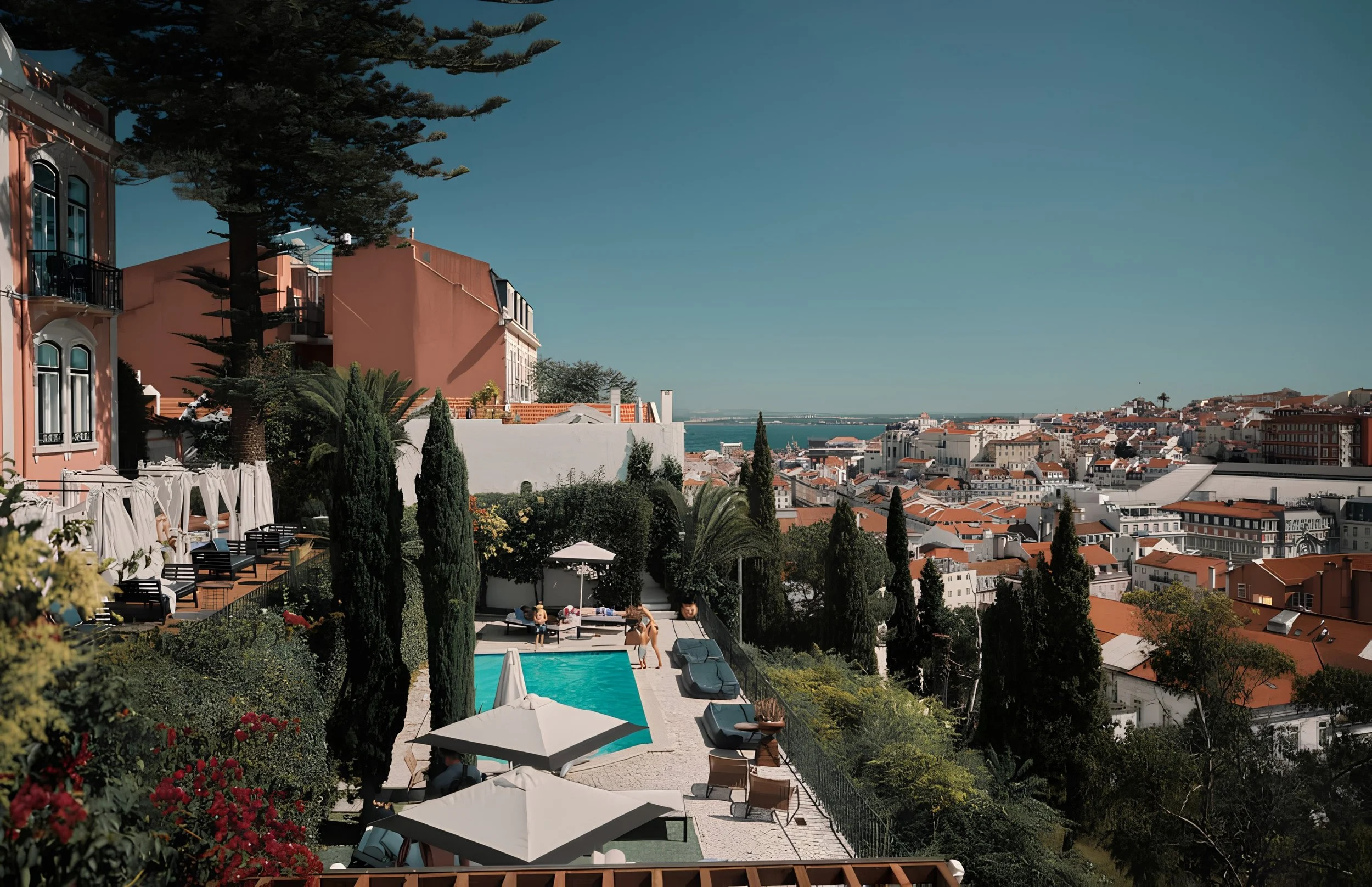 View of a coastal city from a hillside with buildings, trees, a swimming pool, sunbathers, umbrellas, and chairs, overlooking the ocean in the distance.