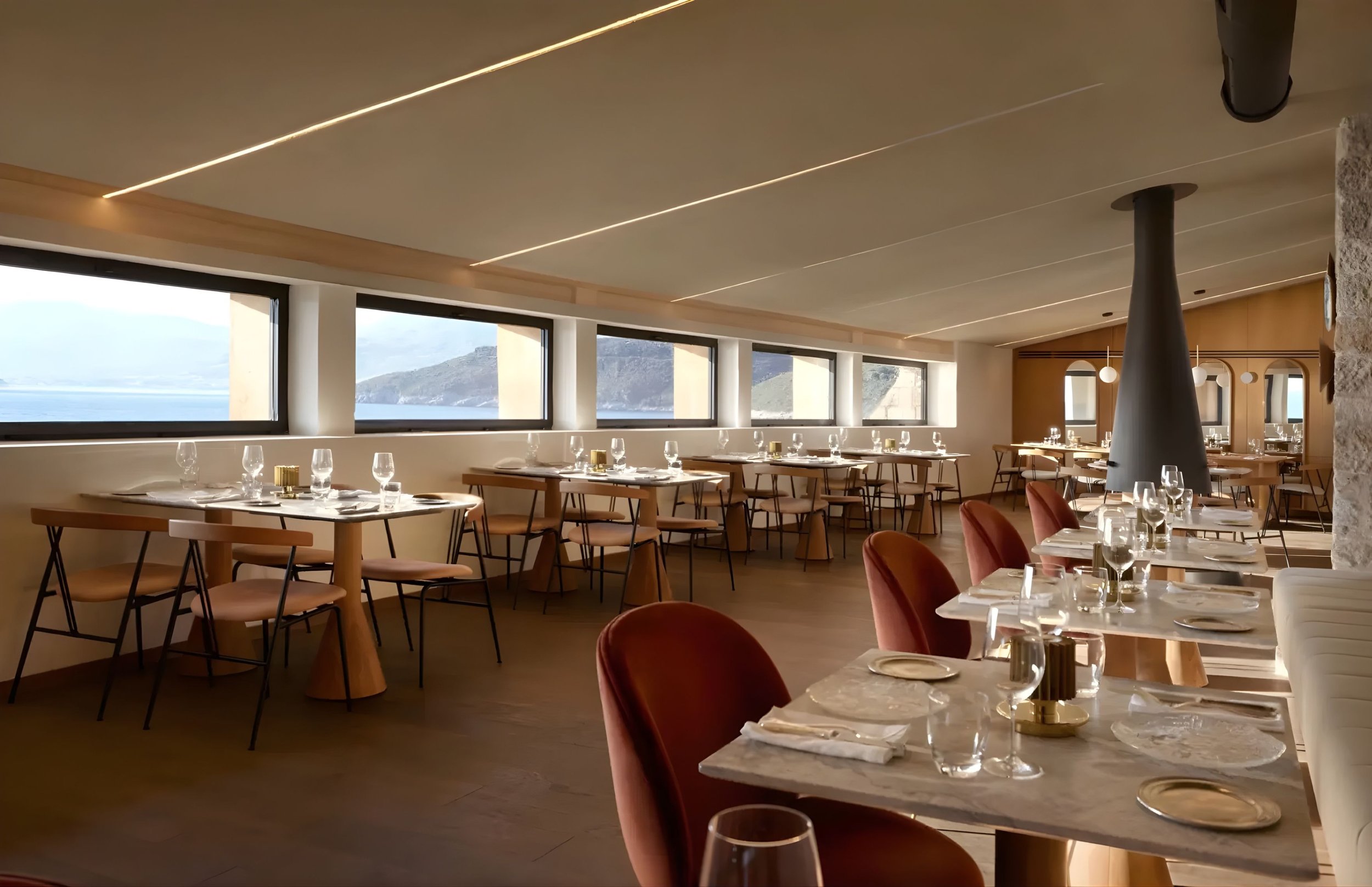Elegant restaurant interior with large windows overlooking mountains and water, set tables with wine glasses, candles, and white tablecloths, modern lighting, and warm decor.