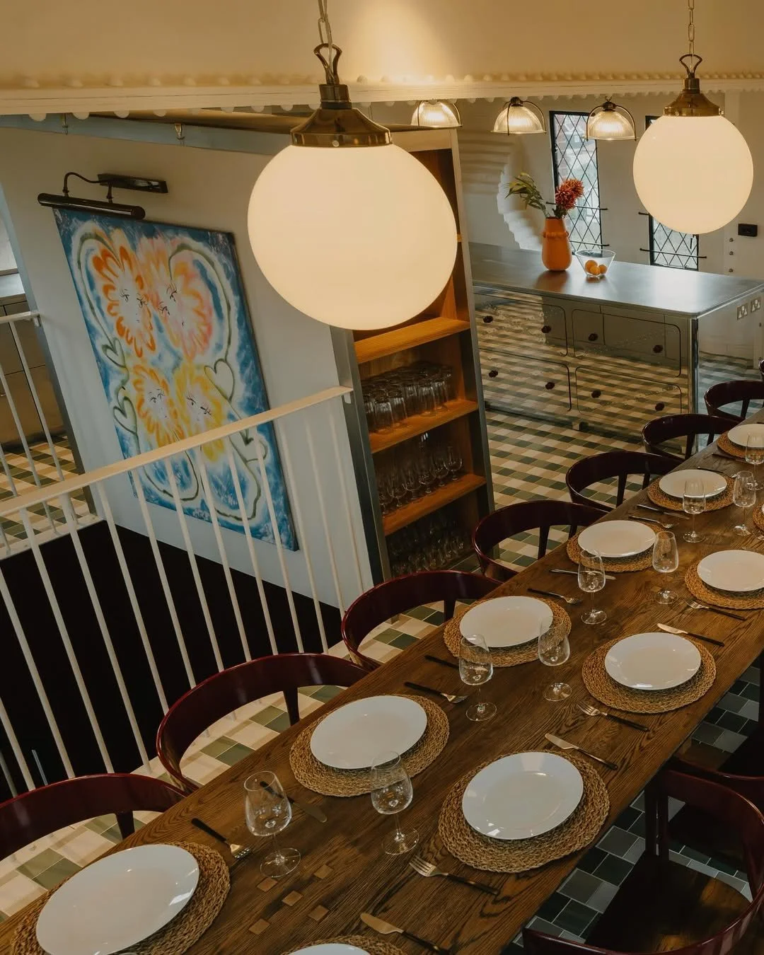 A dining table set with white plates, glassware, forks, knives, and woven placemats, in a well-lit room with large hanging lights and artwork on the wall.