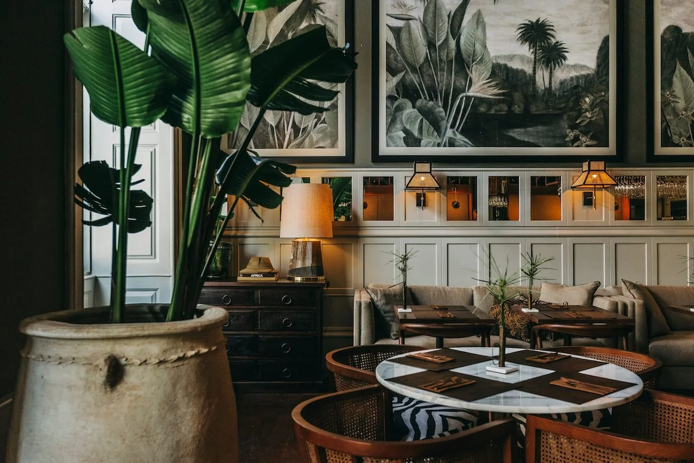 Interior of a cozy restaurant or cafe featuring a large potted plant with broad green leaves in the foreground, a round marble table set for dining in the middle, sofa seating along the wall with small tables, wall art with large green leaves and jungle scenery, warm lighting from lamps and wall fixtures, and a mirror reflecting the warm ambiance.