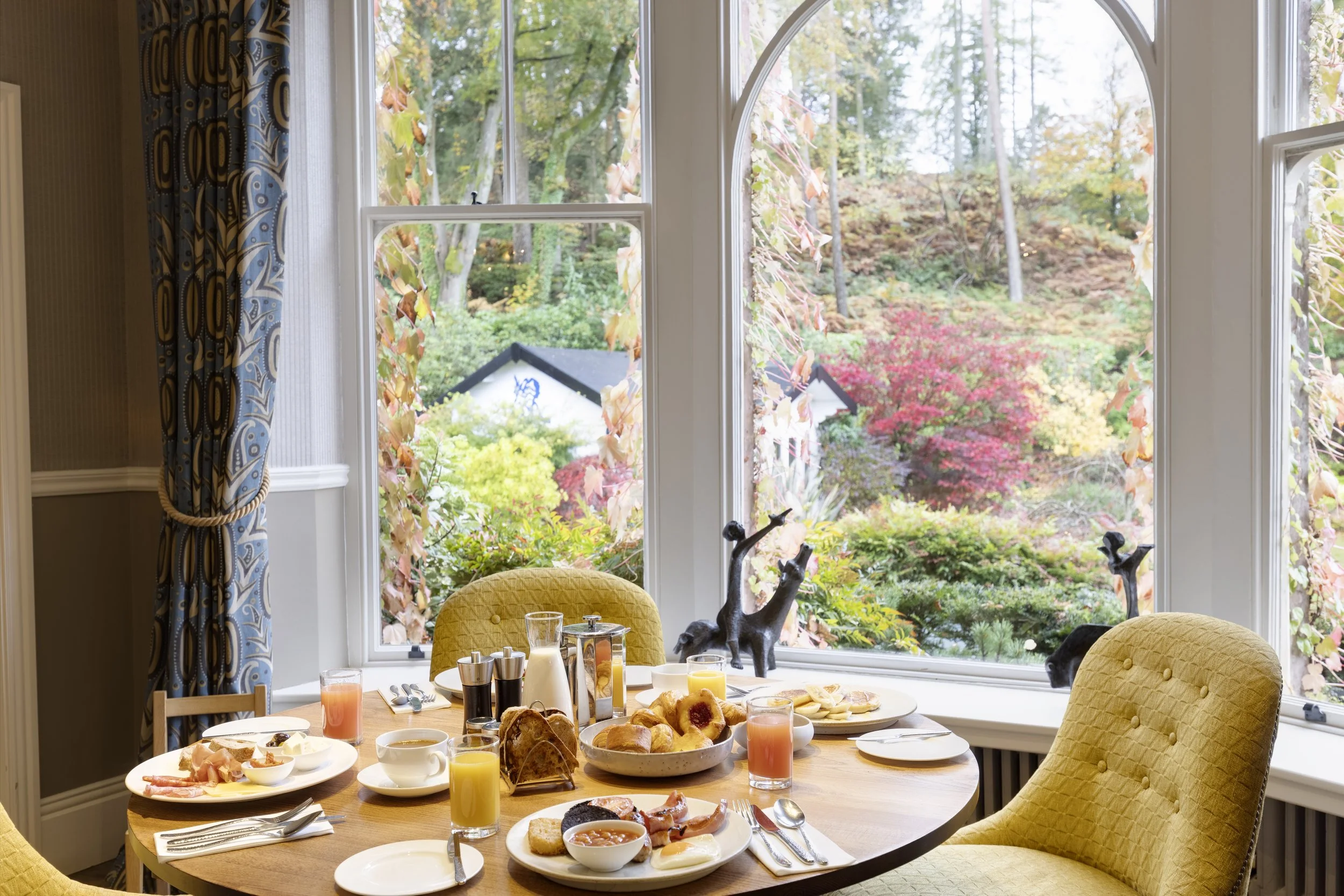 A breakfast table set with various foods and drinks in front of a large window showing trees with autumn leaves. The table has plates of breakfast items, glasses of juice, and utensils. There are yellow upholstered chairs around the table, and the ro