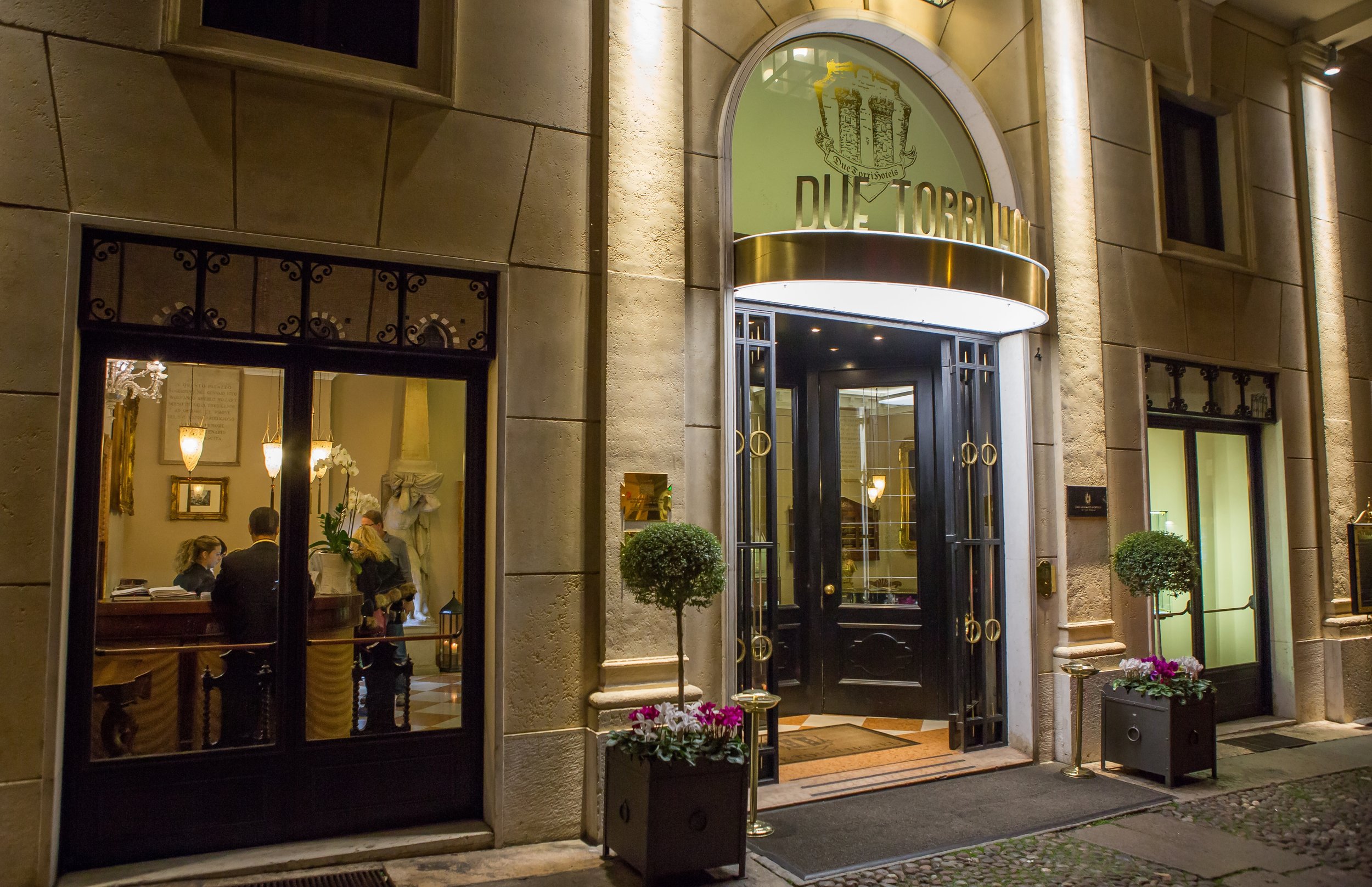 Night view of the entrance to a hotel named Due Torri, with a glass door, potted plants with flowers, and a window showing the lobby with people inside.
