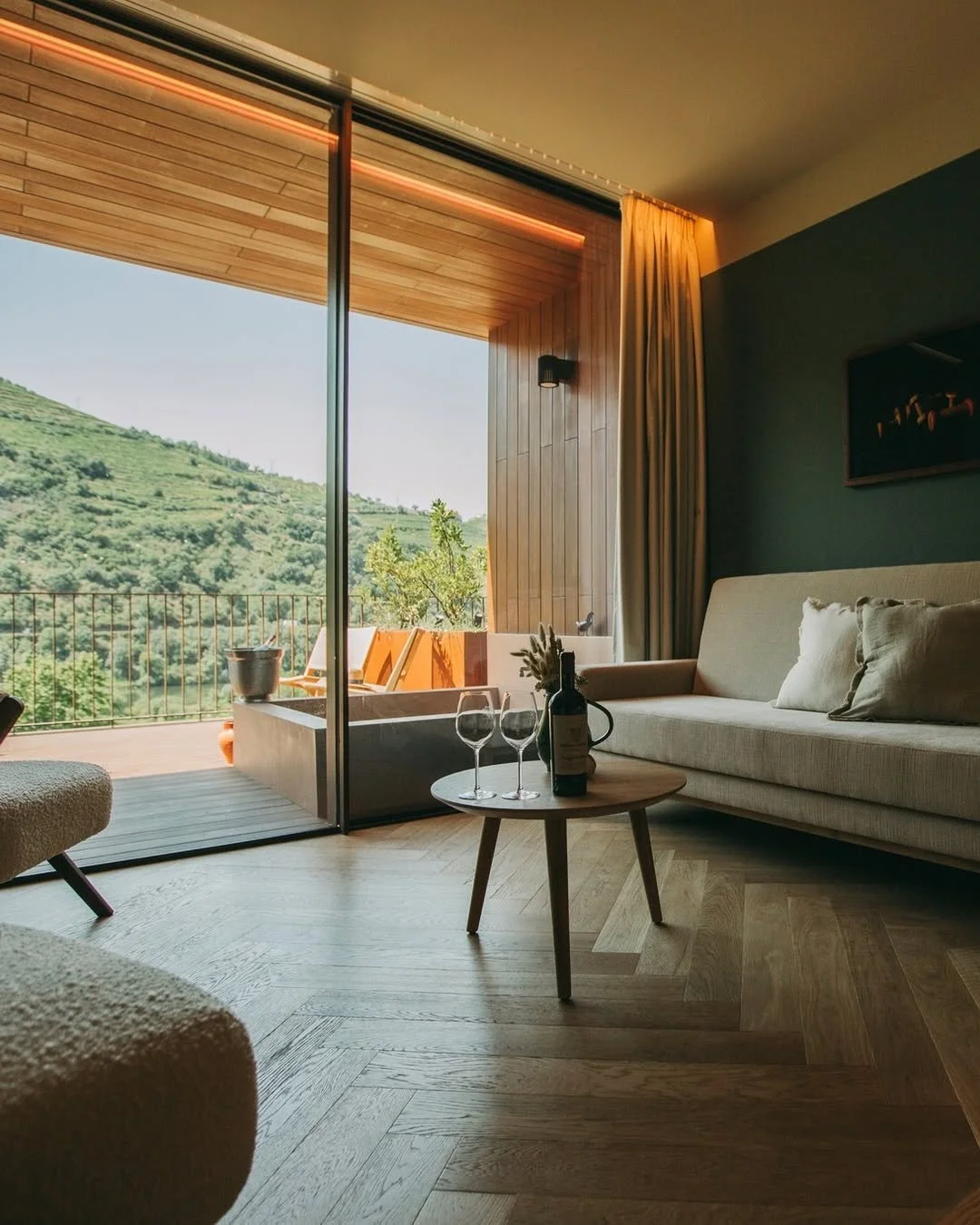 Living room with a view of a green hillside through glass sliding doors, furnished with a beige sofa, a small round wooden table with wine bottle and glasses, and a balcony outside with wooden deck chairs.