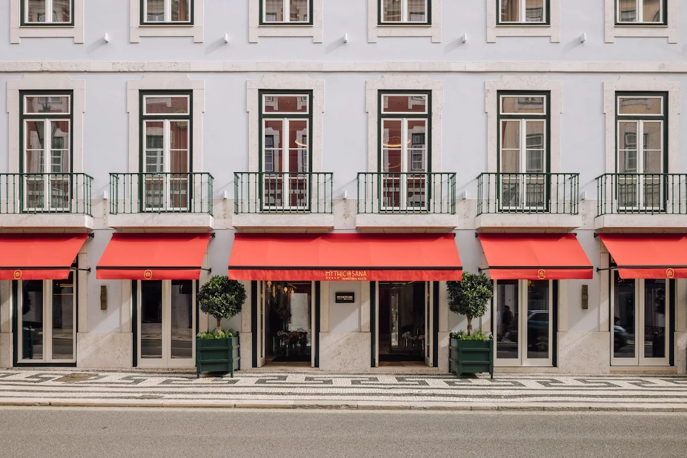 Facade of a building with large windows, small balconies, red awnings, and green potted trees outside a restaurant entrance.