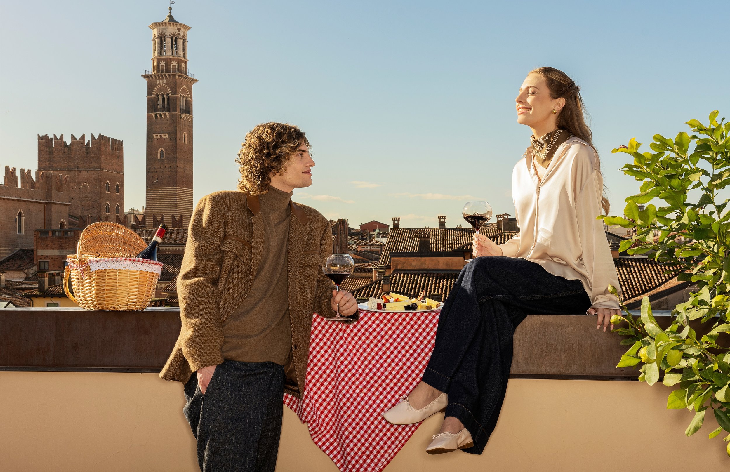 A man and woman enjoying wine and cheese on a rooftop terrace in a European city, with historic brick buildings and a tower in the background, during daylight.