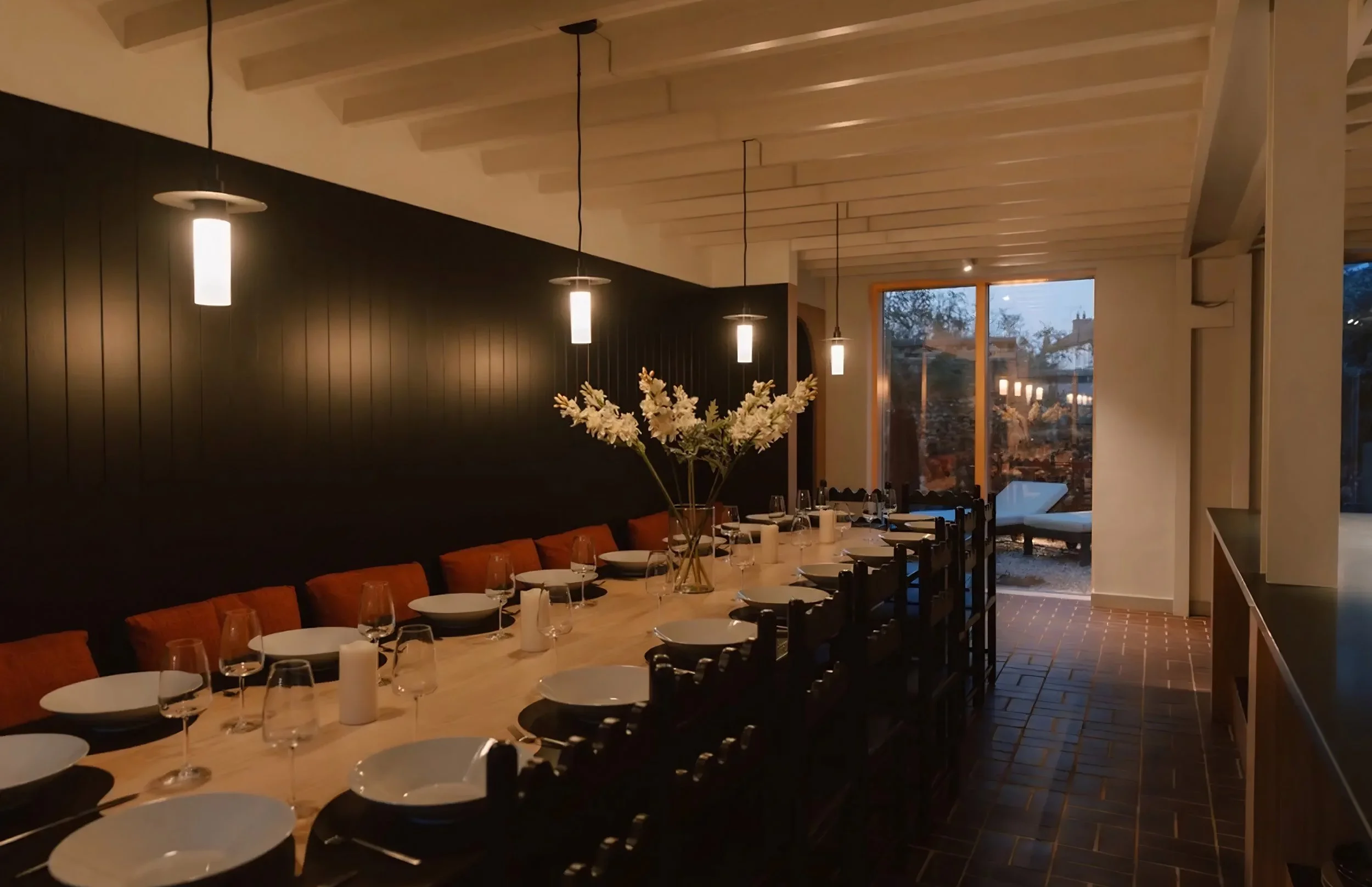 A dining room with a long table set with plates, glasses, and floral centerpiece, featuring a black accent wall with pendant lights and large window showing an outdoor scene during twilight.