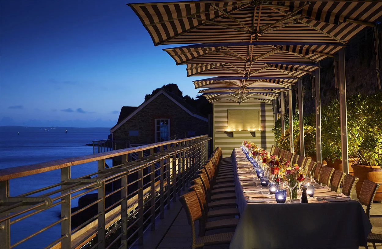 Nighttime outdoor dining area on a balcony overlooking the water, with a long table set with flowers, candles, and place settings under striped umbrellas.