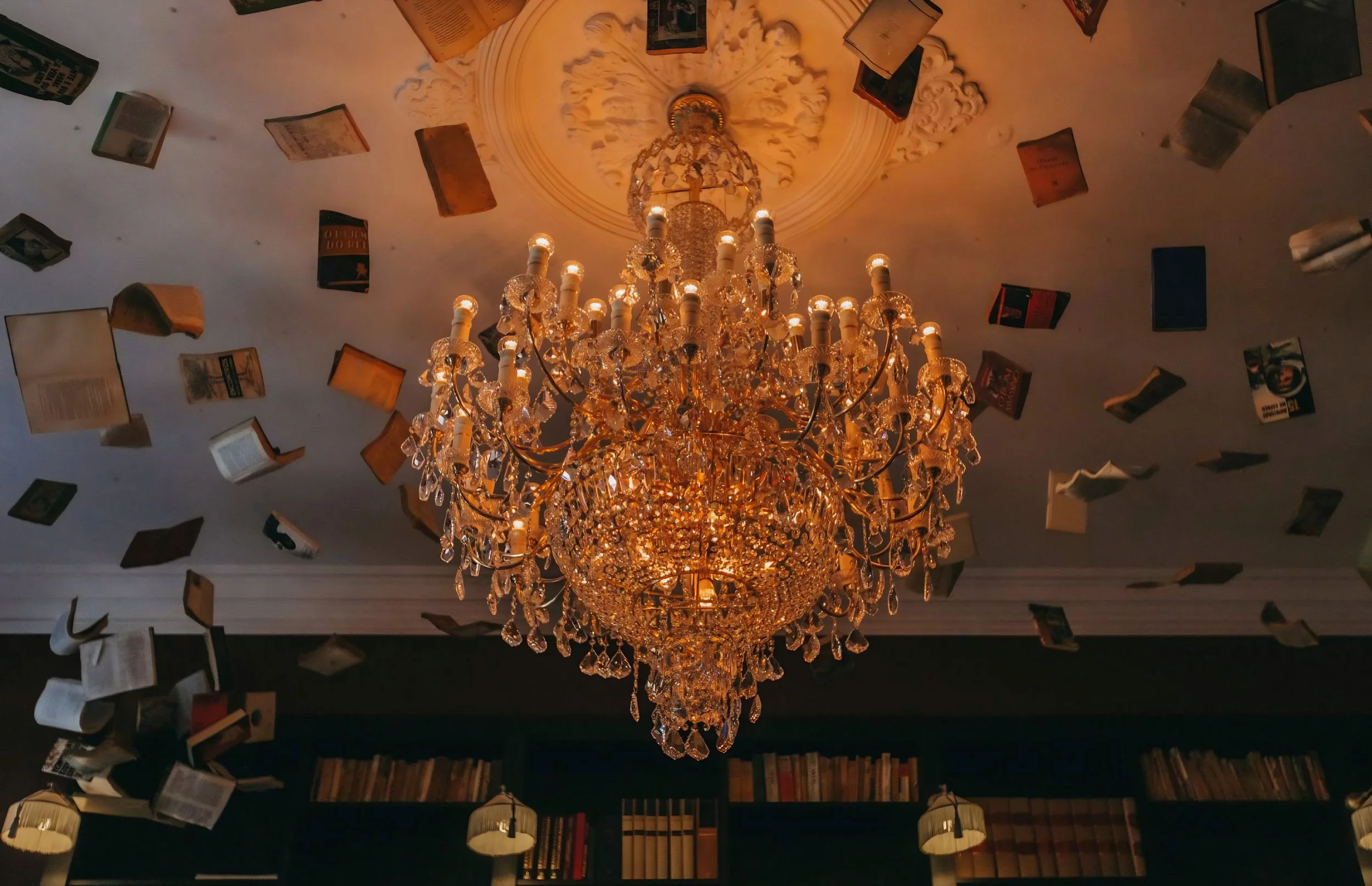 A large ornate chandelier with numerous crystal embellishments hangs from a ceiling. Books and magazines are floating in the air around the chandelier, creating a surreal scene.