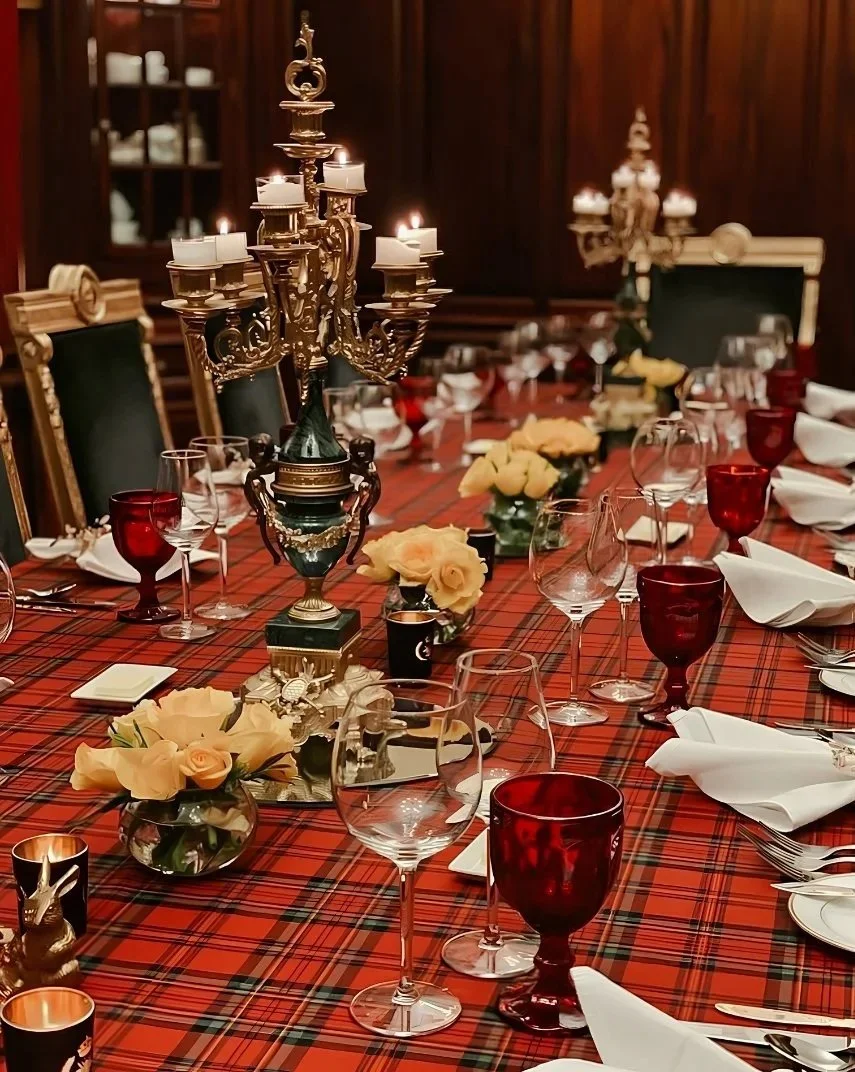 A formal dining table with a red tartan tablecloth, adorned with floral centerpieces, elegant glassware, white napkins, and gold candle holders with lit candles in an ornate setting.