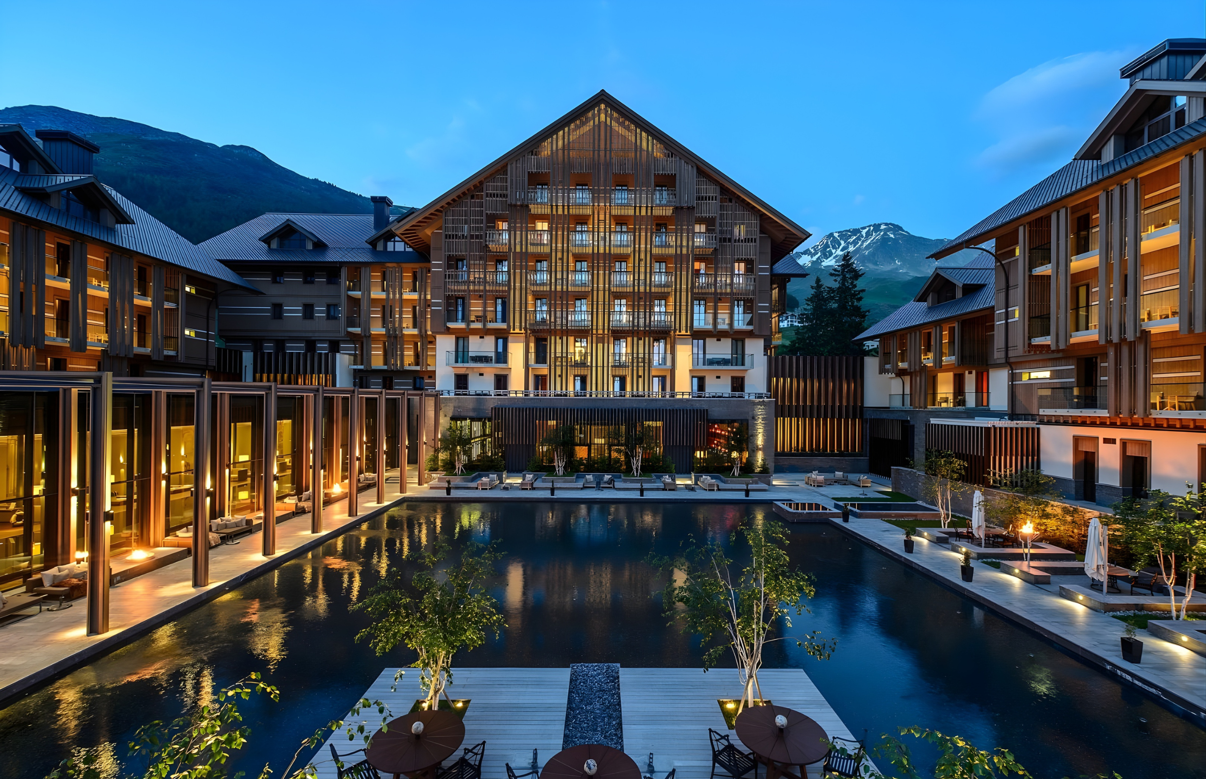A modern hotel or apartment complex at dusk with a central water feature, outdoor seating, and illuminated buildings surrounded by mountains.
