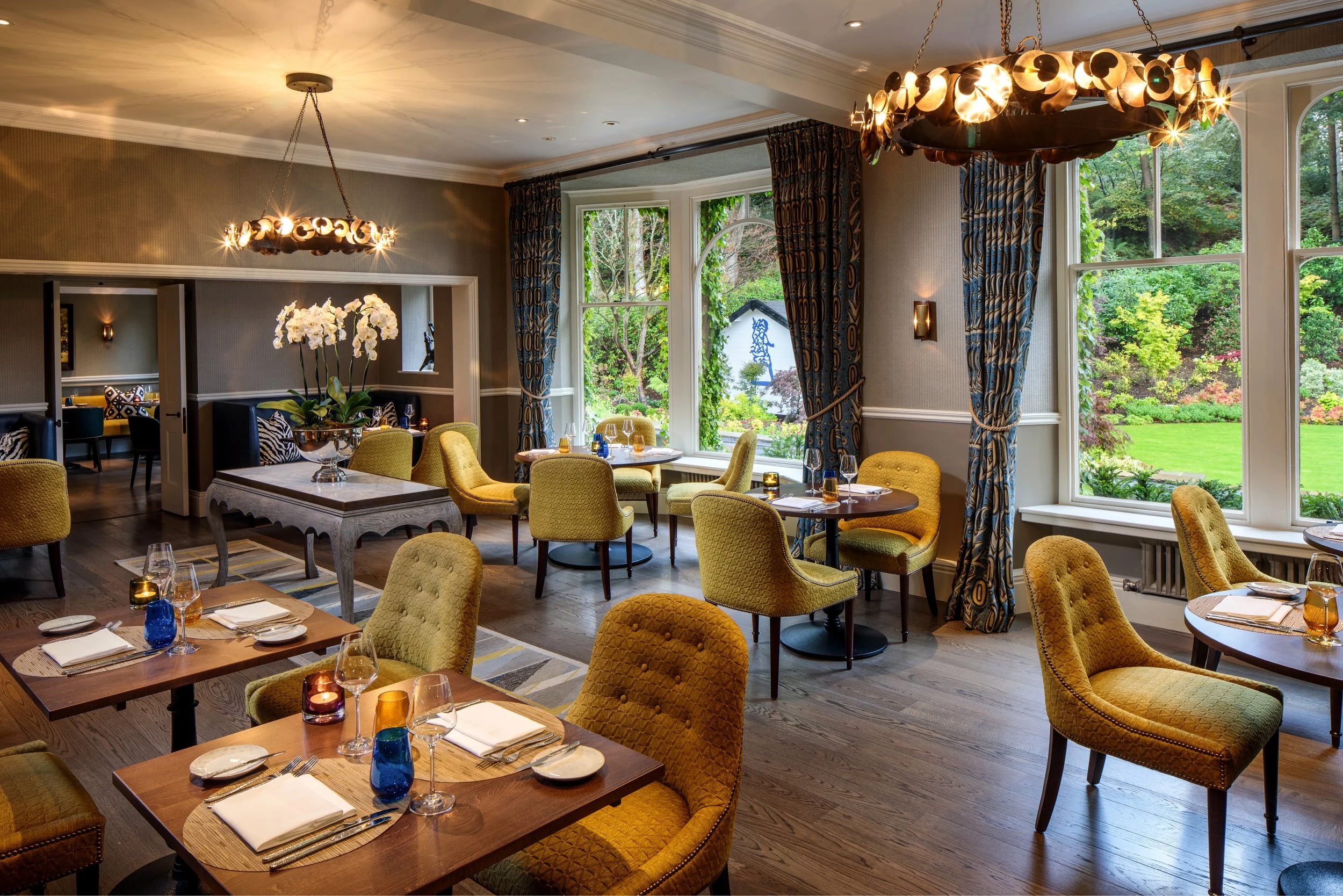 Elegant dining room with yellow upholstered chairs, tables set with glassware and candles, large windows with patterned curtains revealing a lush green garden, and two modern chandeliers hanging from the ceiling.
