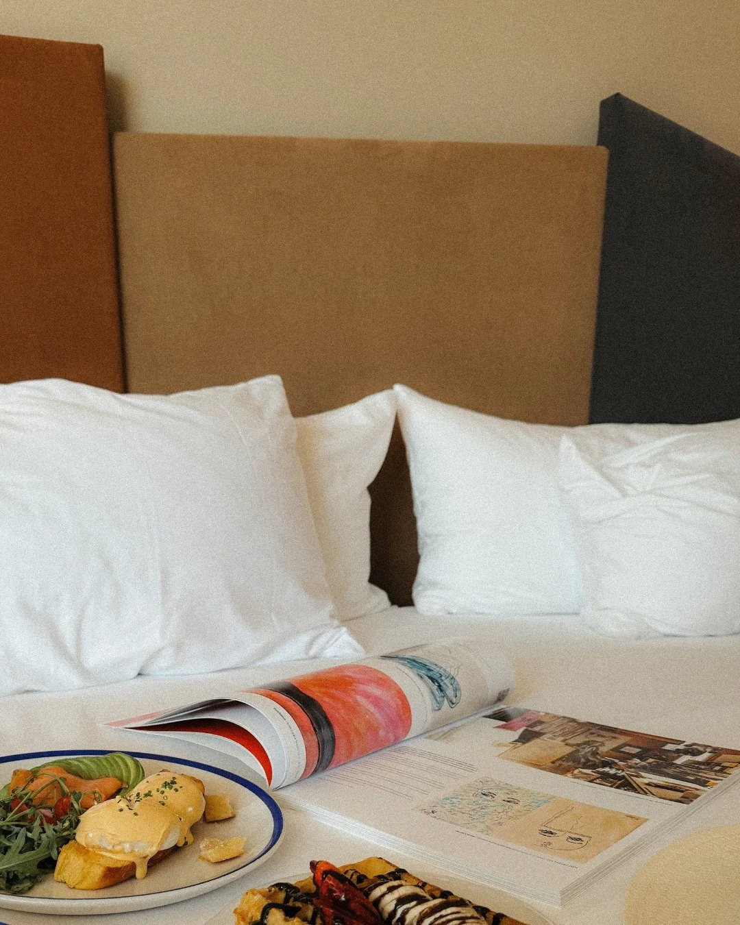A hotel bed with white pillows, a beige headboard, and a magazine on the bed. In front of the bed, there's a breakfast plate with eggs benedict, vegetables, and desserts with toppings.