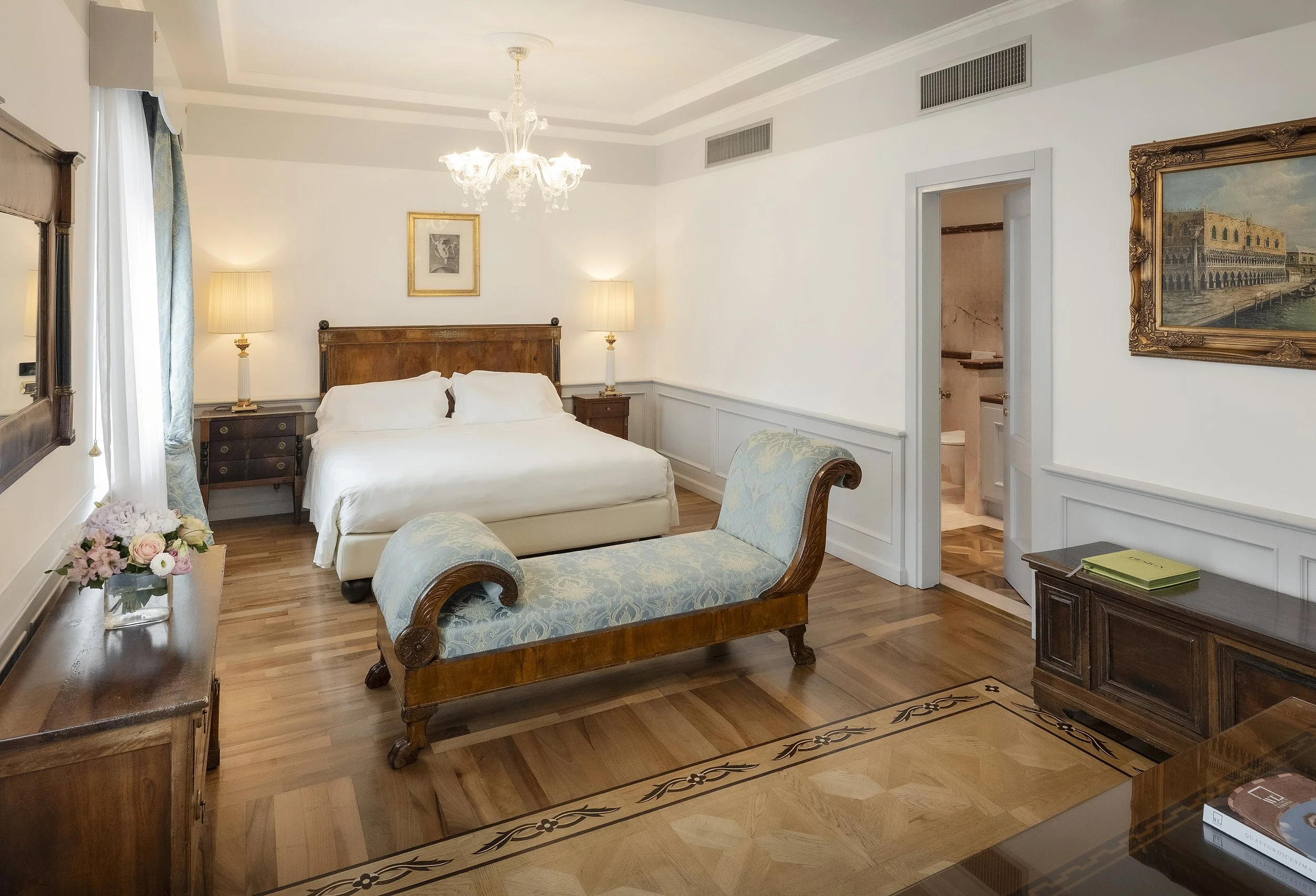 A hotel room with a large bed, side tables with lamps, a floral armchair, a mirror, and framed artwork on the walls. There is a wooden dresser with a flower arrangement and a window with curtains. The room has hardwood flooring and a chandelier hanging from the ceiling. An open door reveals a bathroom.
