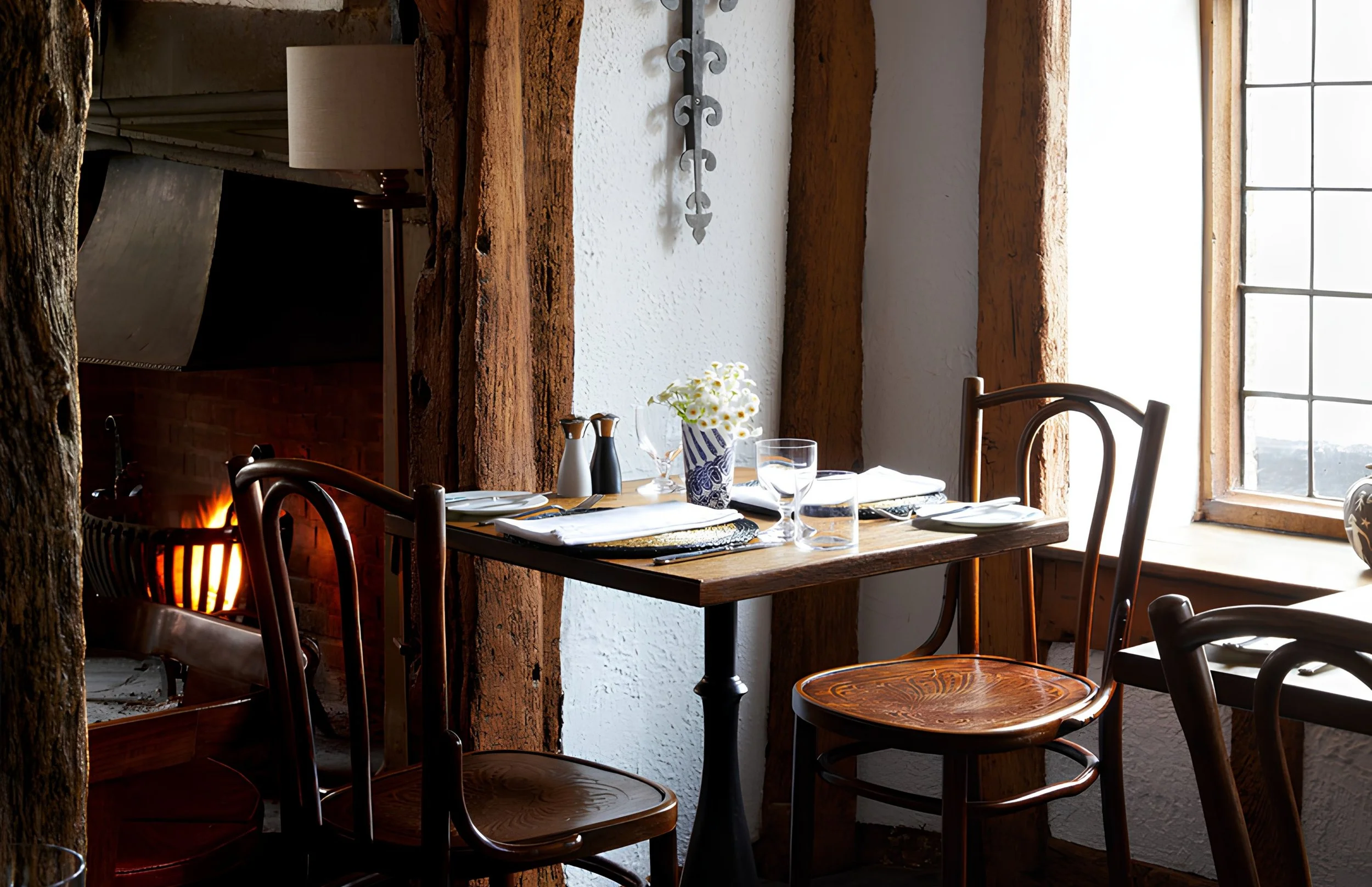 Dining table set with plates, glasses, cutlery, and a flower vase in a rustic restaurant with wooden beams and large windows.
