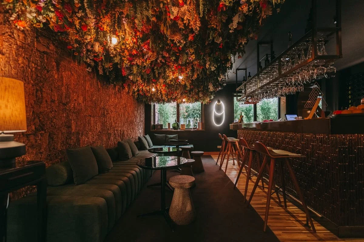 A cozy bar with a dark and warm ambiance, featuring a ceiling decorated with an abundance of flowers and foliage, a brick wall, a green sofa with throw pillows, small round tables, bar stools at the counter, and a neon light on the wall, with large windows showing greenery outside.