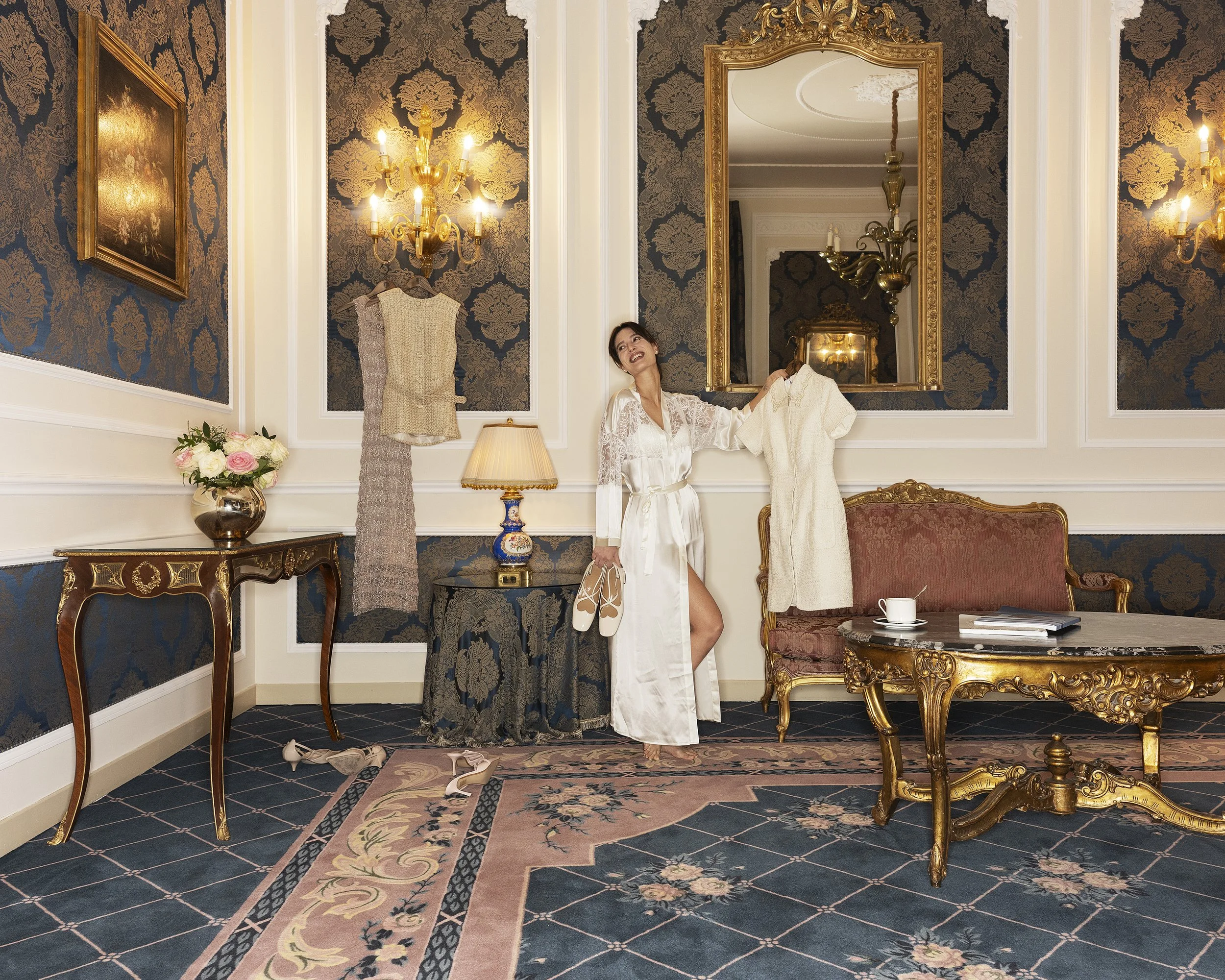 A woman in a white lace dress is holding a hanger with a dress on it, smiling. The room appears luxurious with dark patterned wallpaper, ornate gold-framed mirror, chandelier, matching wall sconces, floral arrangement, vintage furniture, and a decora