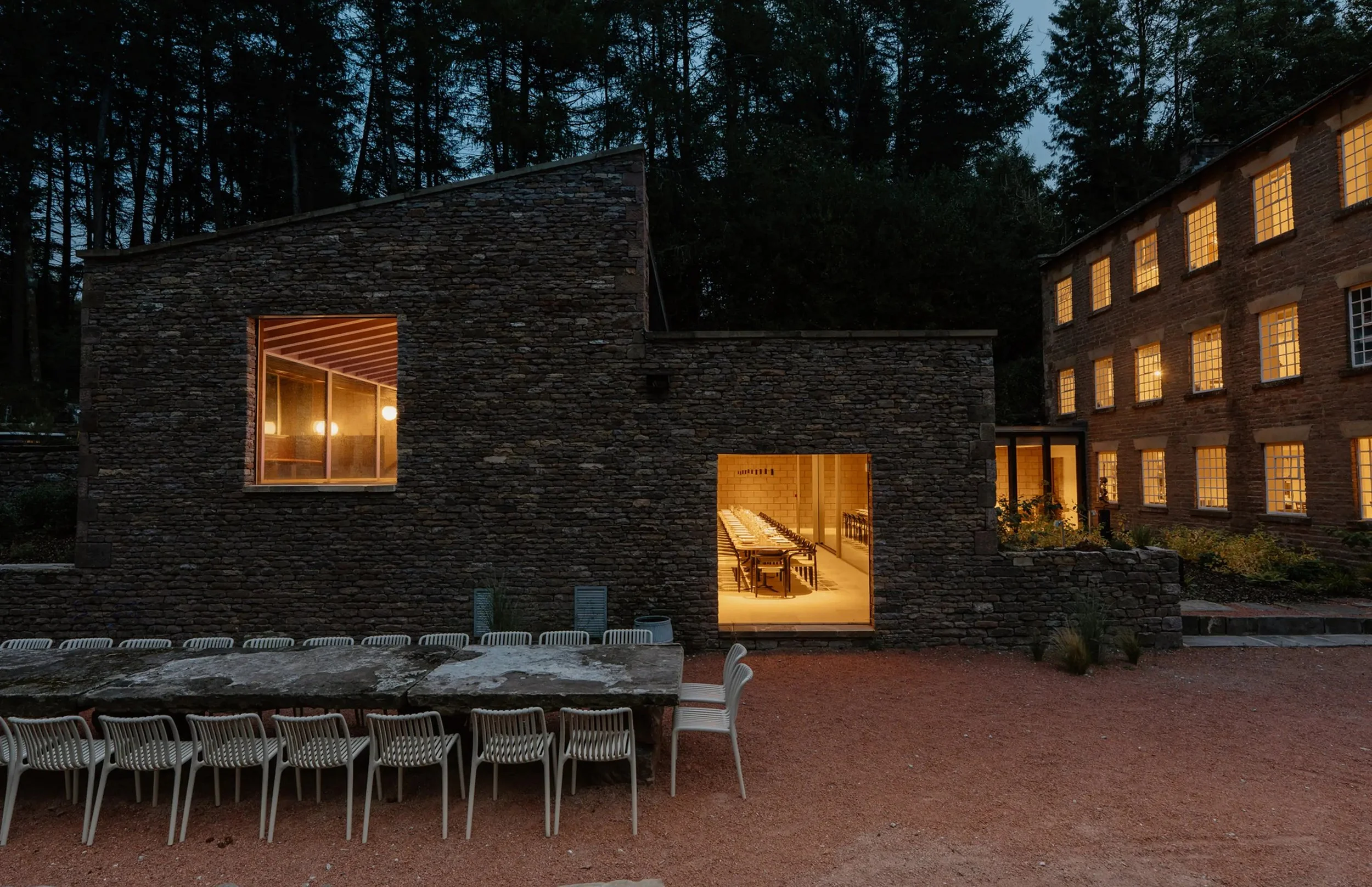 Exterior of a modern stone and brick building at dusk with warm indoor lighting and an outdoor patio with white chairs and a stone table.