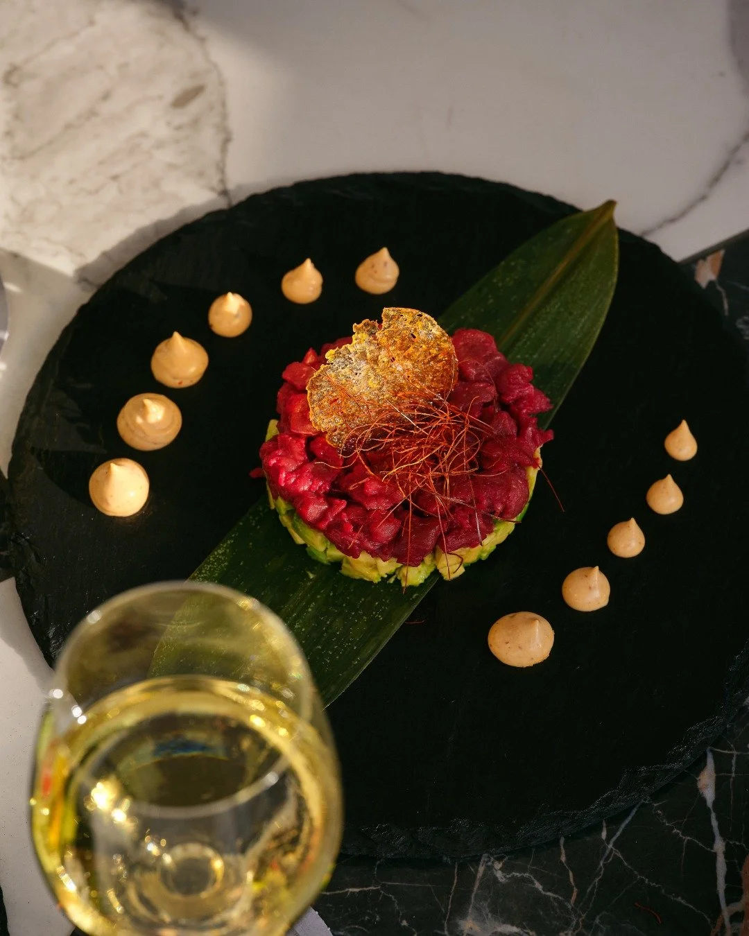 Elegant dish featuring diced raw tuna on a bed of shredded avocado, garnished with a crispy leaf and fine red threads, served on a dark plate with evenly spaced dollops of sauce and a glass of champagne nearby.
