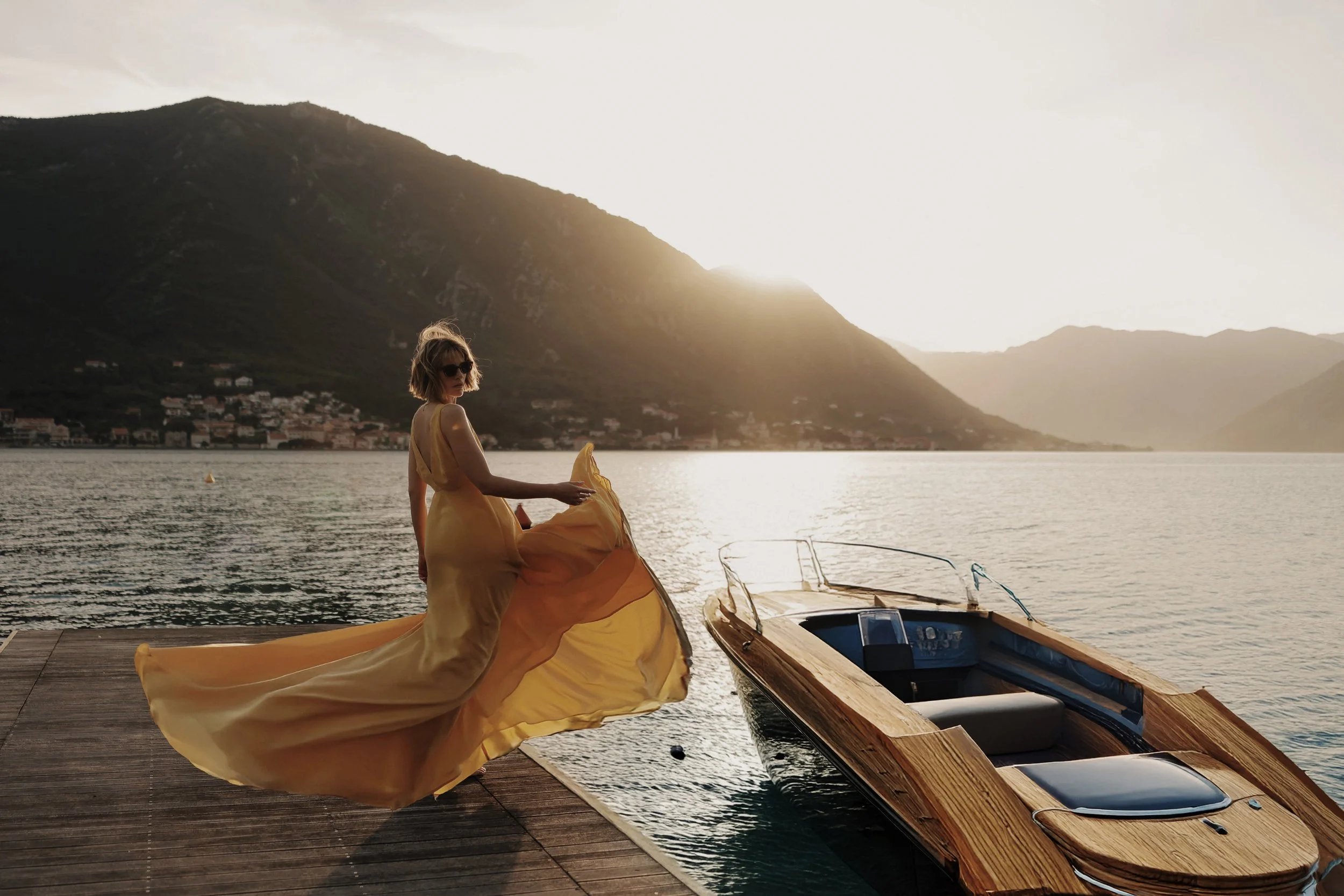 A woman in a flowing yellow dress and sunglasses stands on a dock by the water at sunset, with mountains in the background and a small wooden boat nearby.