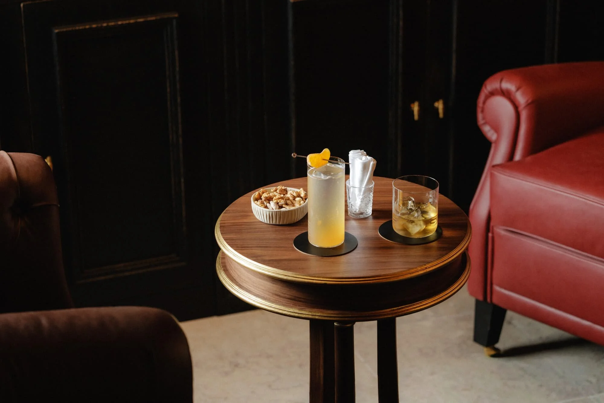 A round wooden table with three beverages, including a cocktail with a yellow garnish, a glass of golden liquid with ice, and another glass of an amber-colored drink. Also on the table is a small bowl of mixed nuts and a clear glass with crumpled nap