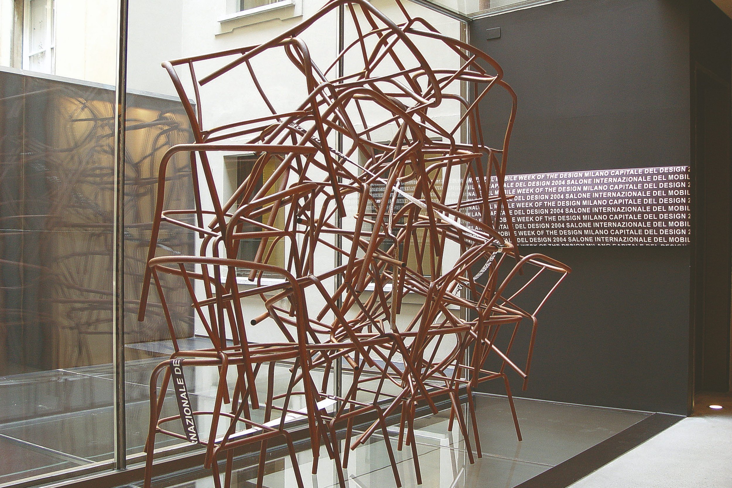 A sculpture of overlapping reddish-brown metal chairs stacked haphazardly inside a building near a large window.