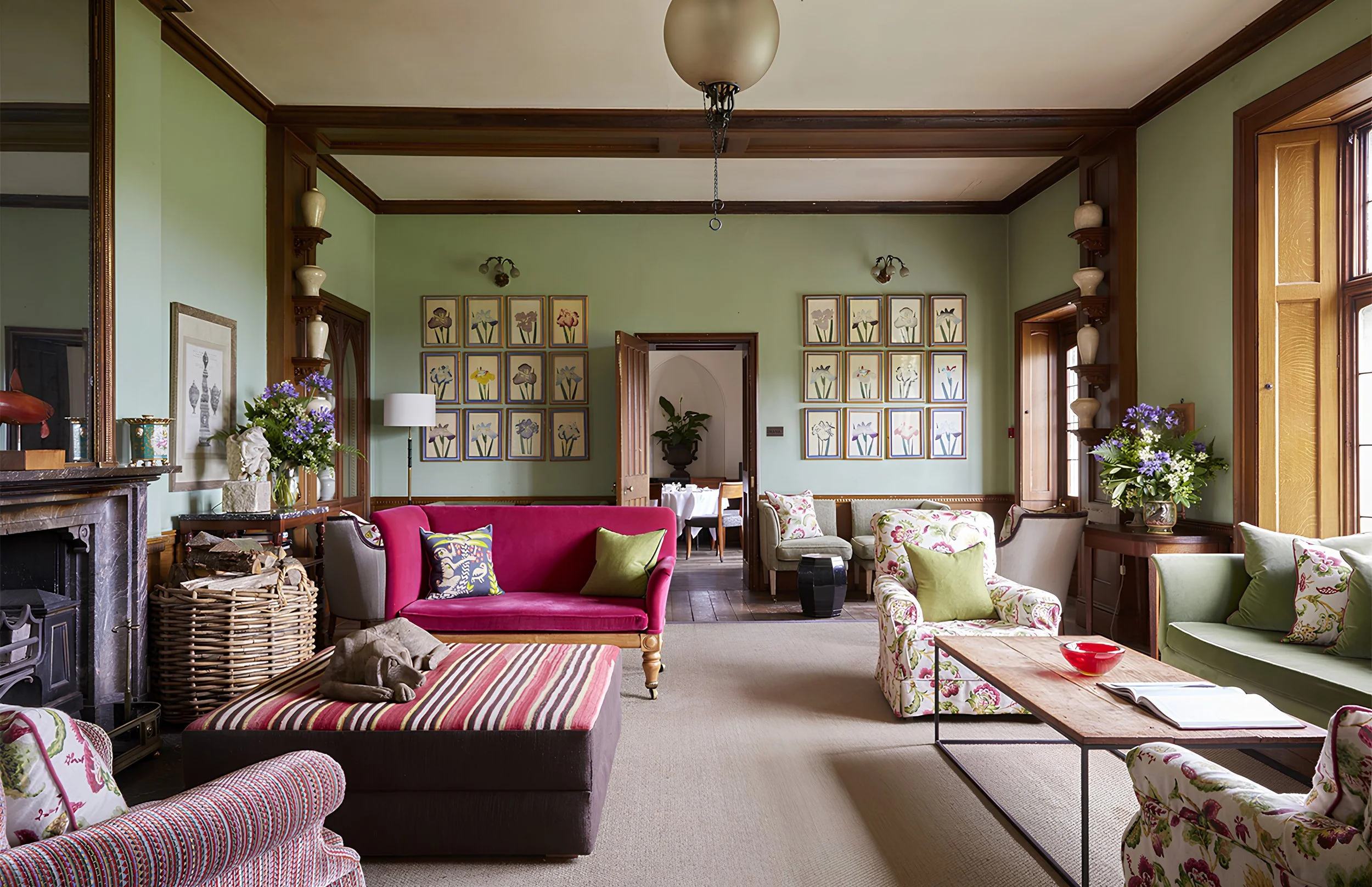 A spacious living room with green walls, large windows, and wooden accents. It features a various colorful sofas and armchairs, floral patterned pillows, and decorative vases, with artwork displaying floral designs on the walls.