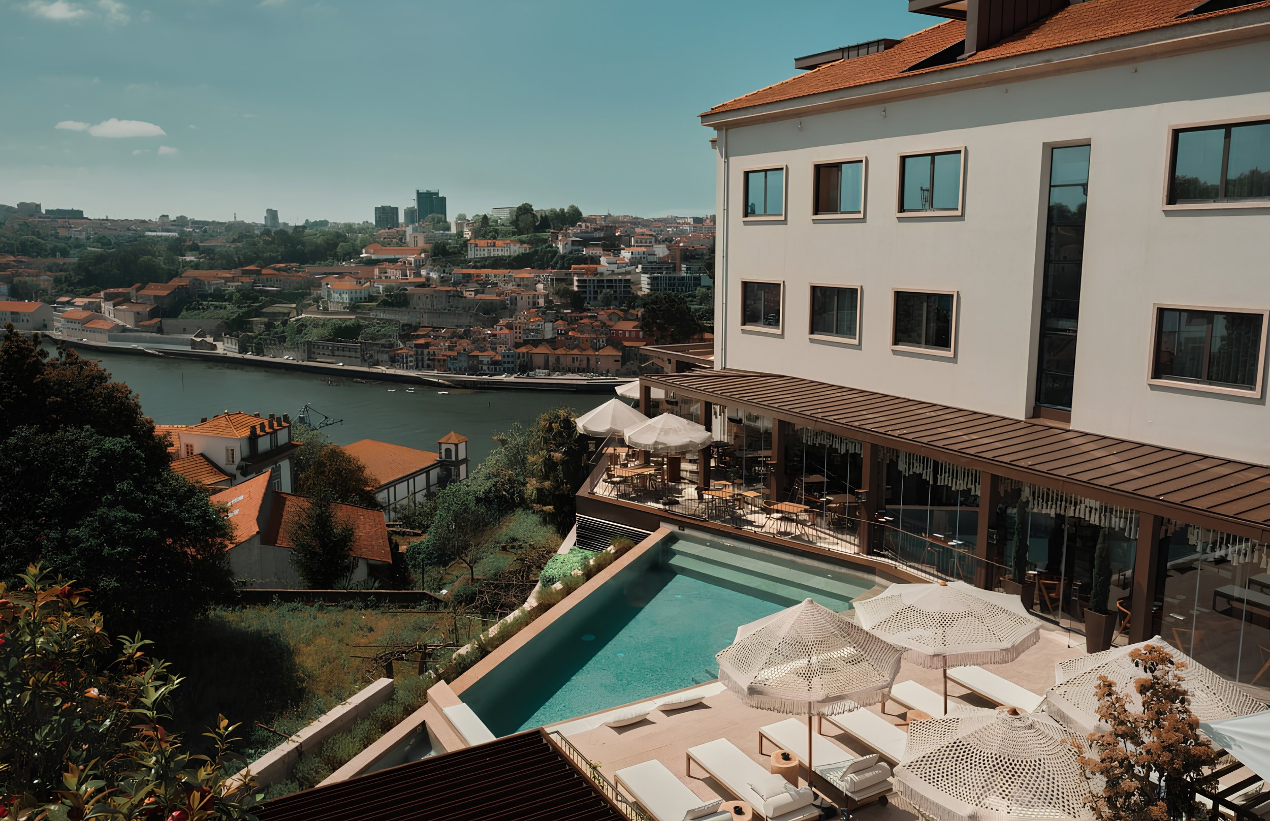 Cityscape view from a balcony featuring a modern building with multiple windows, outdoor seating with umbrellas, a swimming pool, and a river, with a hilly city skyline in the background under blue skies.