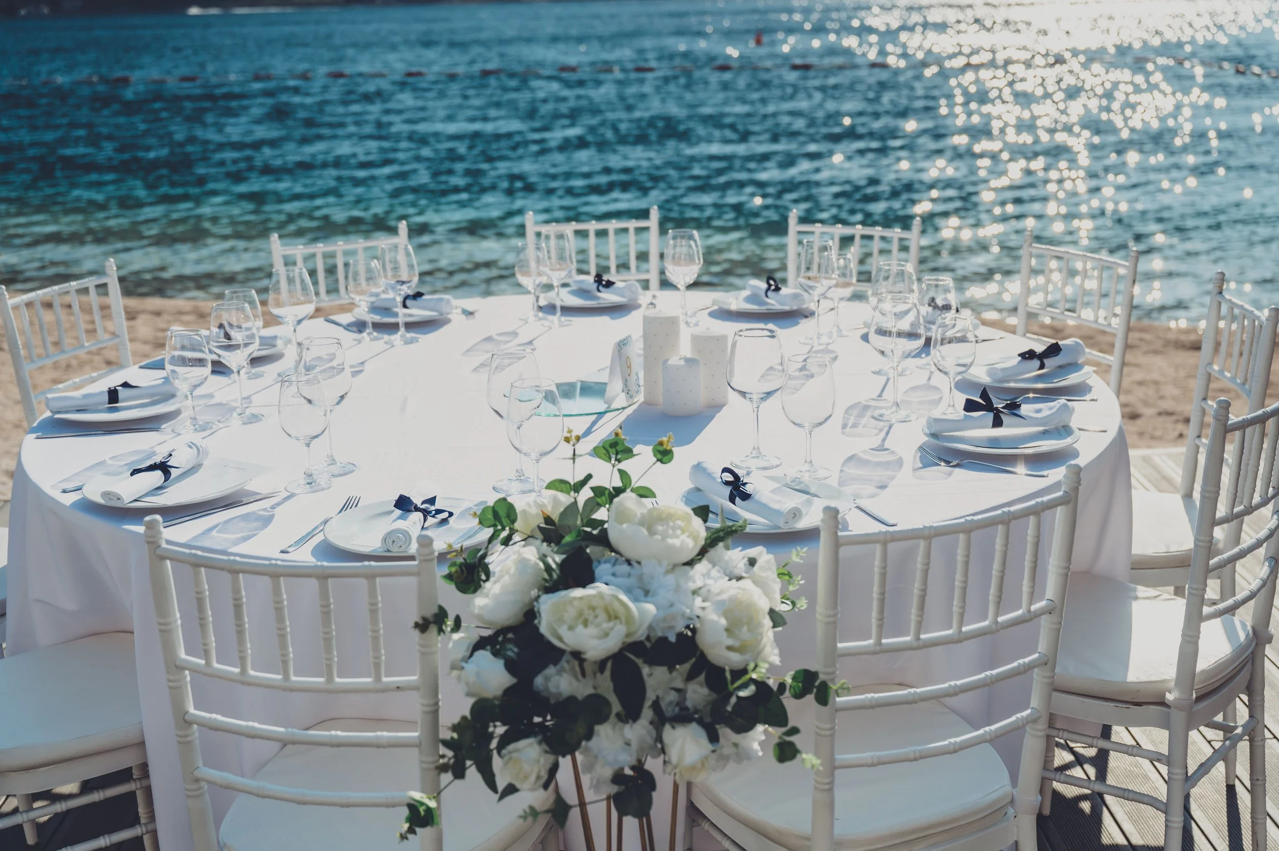 A round table set for a formal event with white tablecloth, wine glasses, and napkins with black ribbons, decorated with a floral centerpiece, by the water with chairs around, overlooking a sunny ocean.