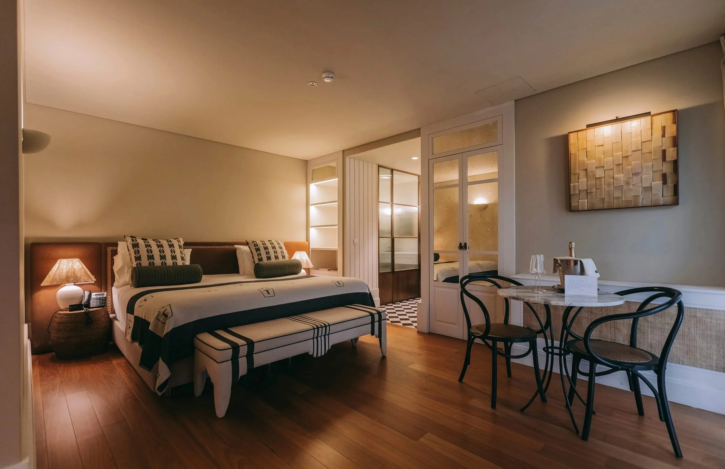 View of a hotel room with a bed, nightstand, lamps, a small dining table with chairs, and a decorative wall art.