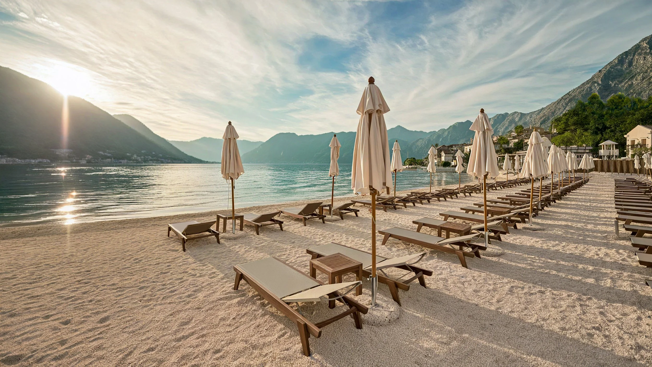 A peaceful beach scene with numerous empty lounge chairs and closed umbrellas on sandy shore, calm water reflecting the sky, mountains in the background, and the sun rising over the hills.