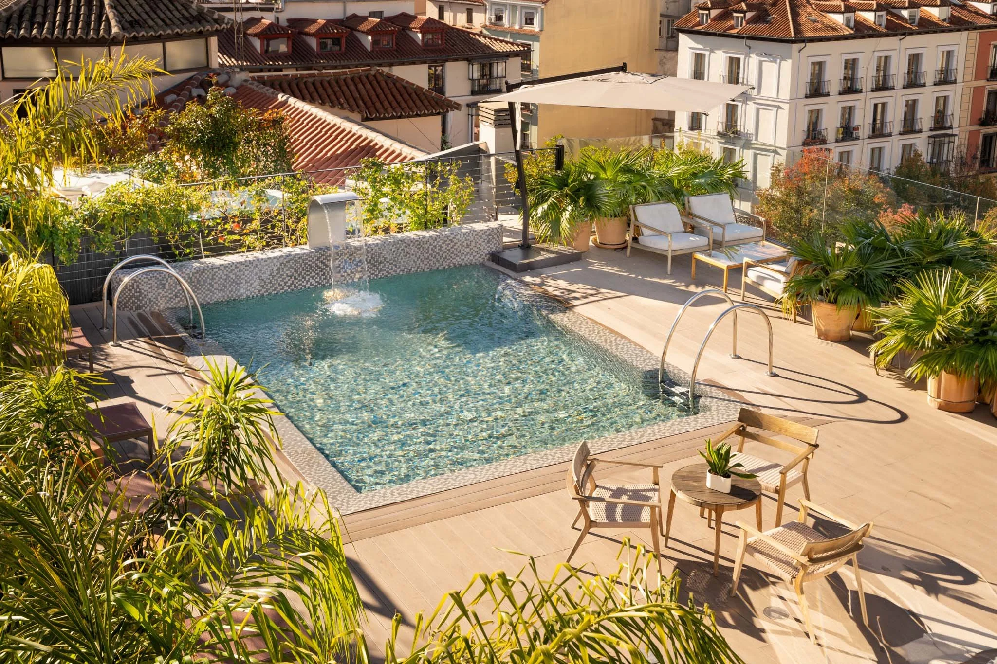 Rooftop swimming pool with outdoor seating and lush green plants, surrounded by city buildings with red-tiled roofs in warm sunlight.