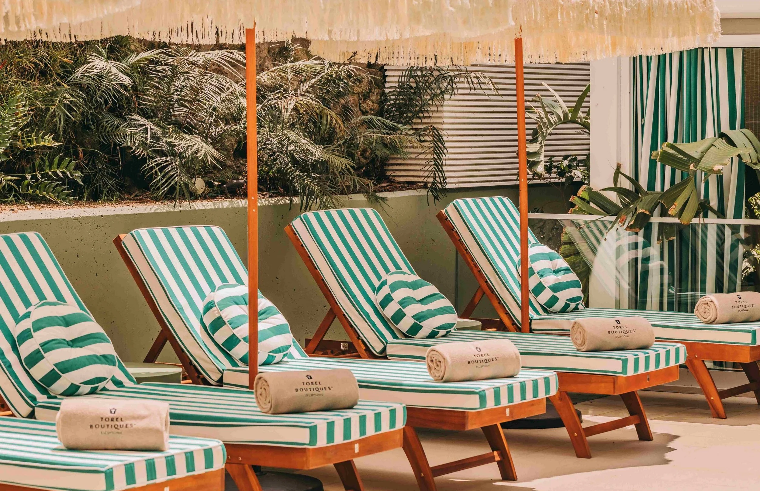 Poolside chairs with green and white striped cushions, rolled towels, and umbrellas on a terrace with lush greenery in the background.
