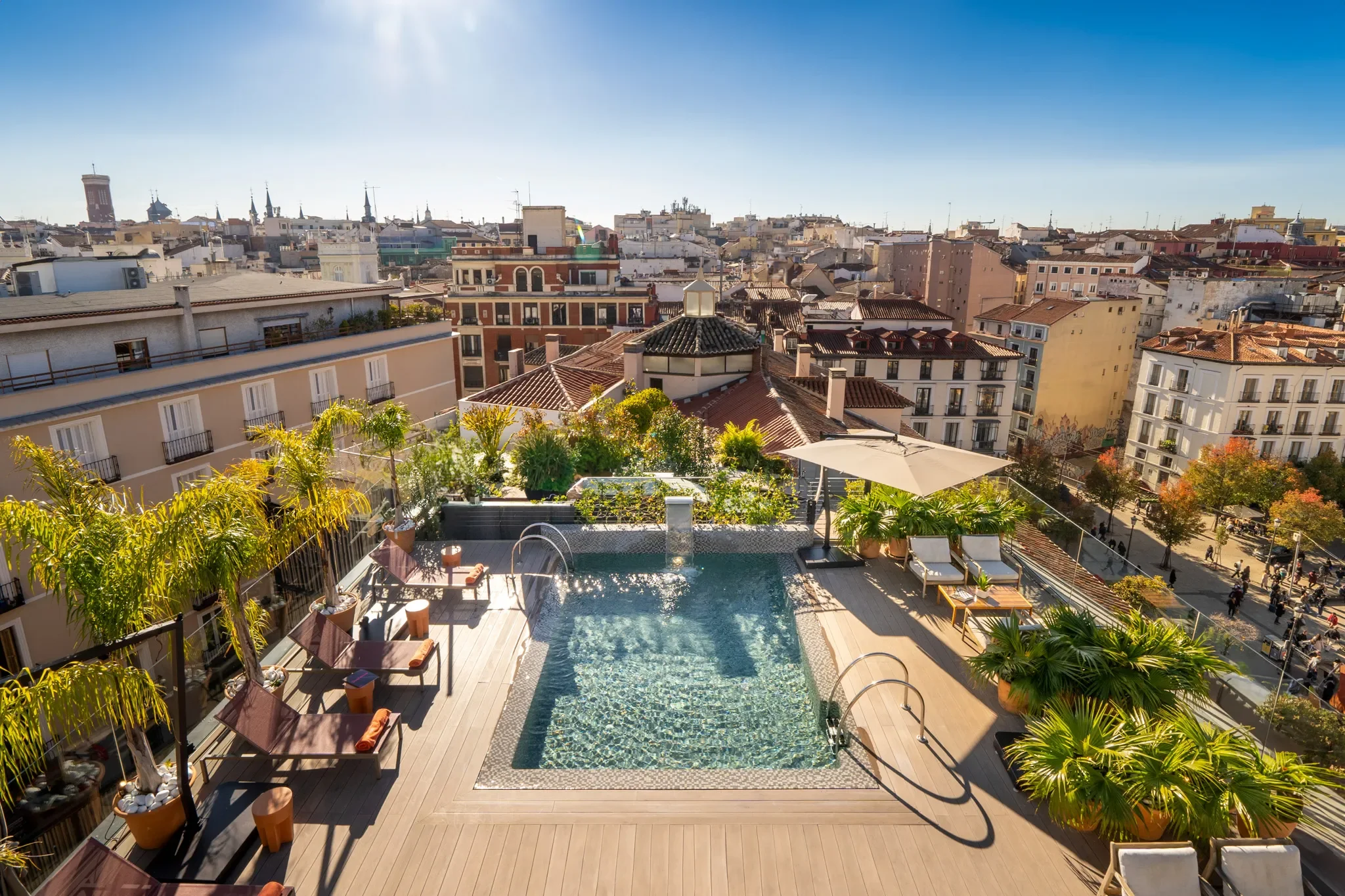 View of a rooftop swimming pool area with lounge chairs and umbrellas, overlooking a city skyline with many buildings and a bright blue sky.