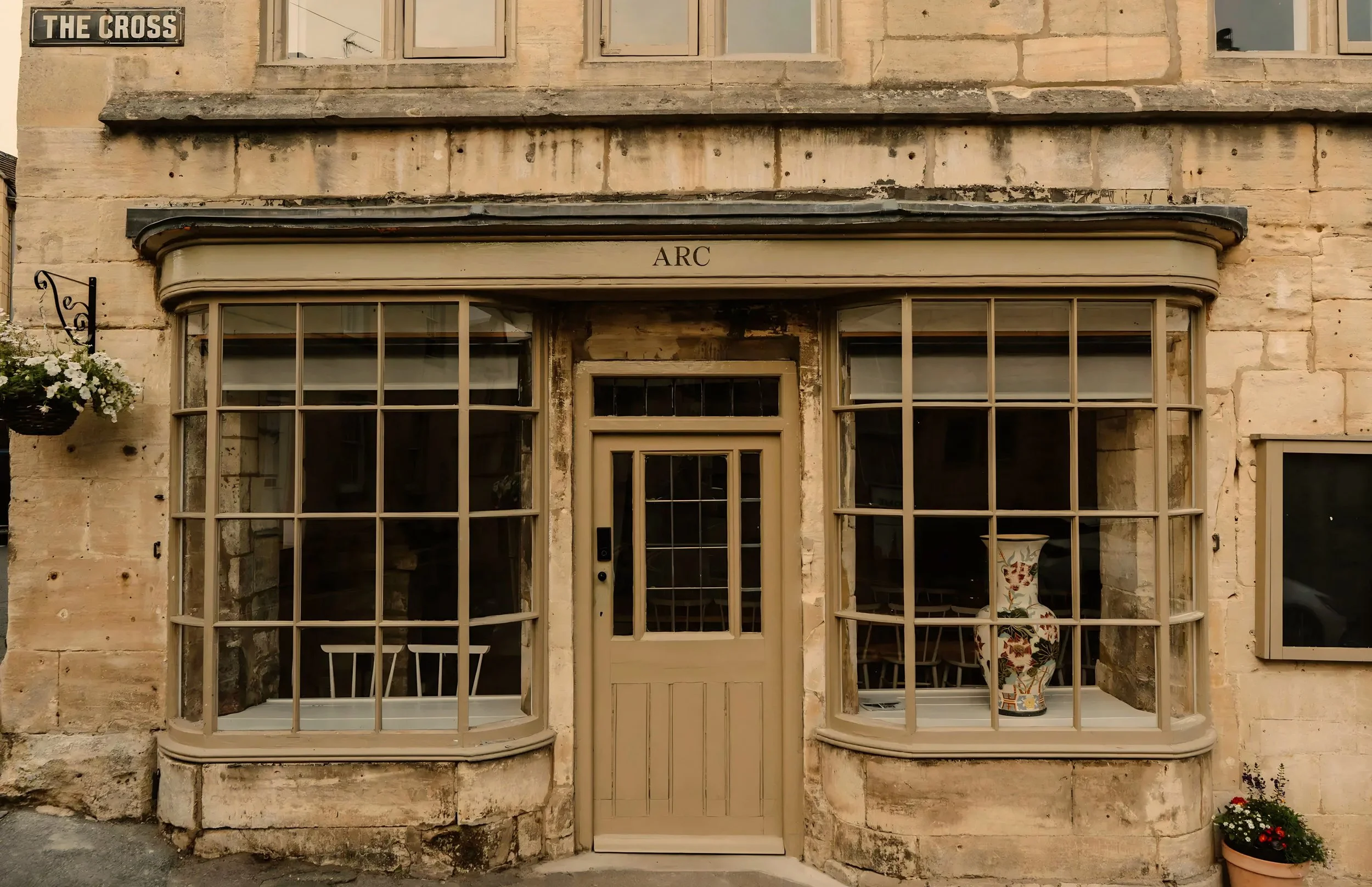 The entrance of a building with a vintage window display, a wooden door, and a sign that says 'The Cross'. There are potted flowers on the side.