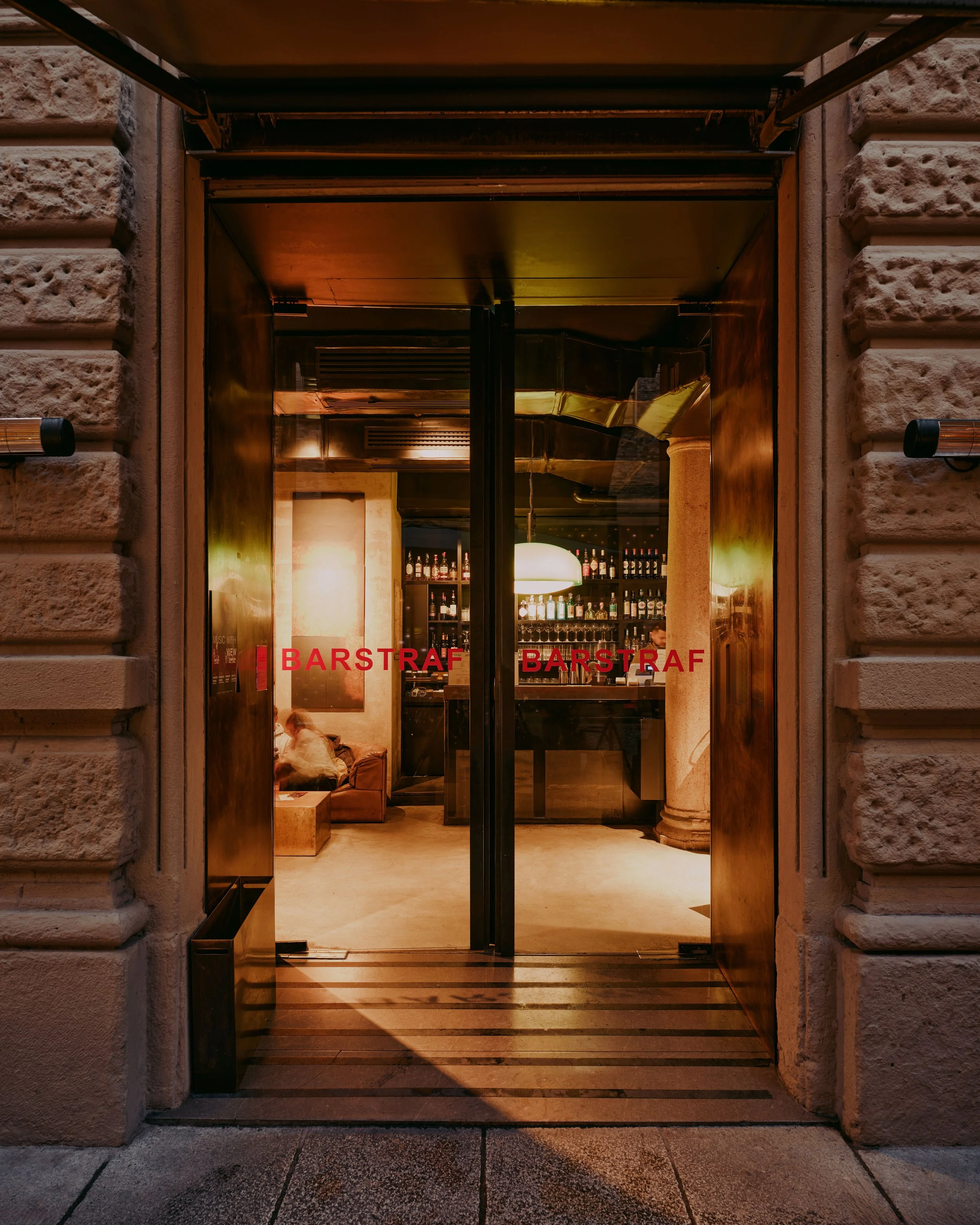 View through glass double doors into a warmly lit bar interior with shelves of bottles, seating, and a hanging light