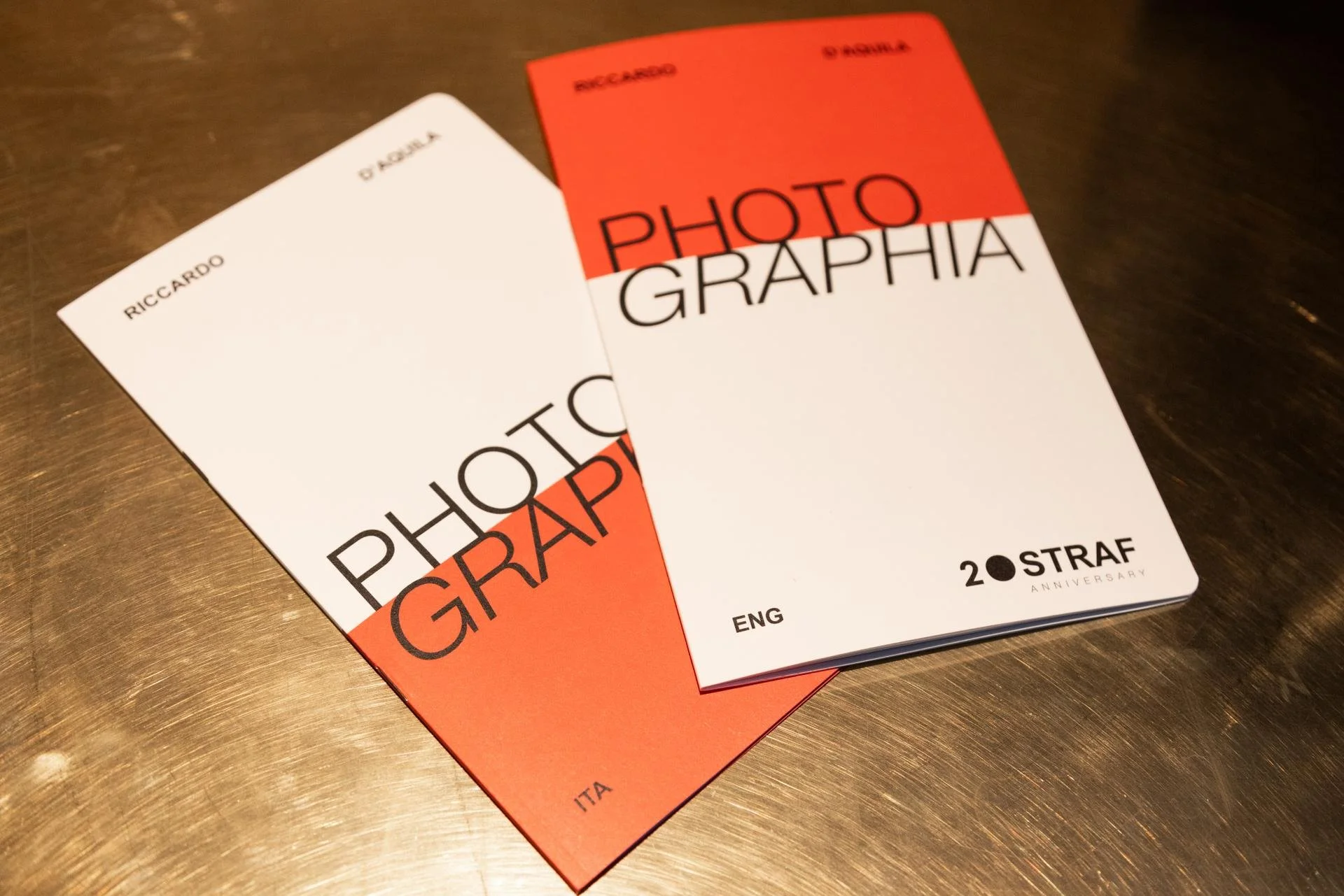 Two books titled 'Photographography' by Riccardo, one with a red and white cover and the other with a white cover, placed on a wooden surface.